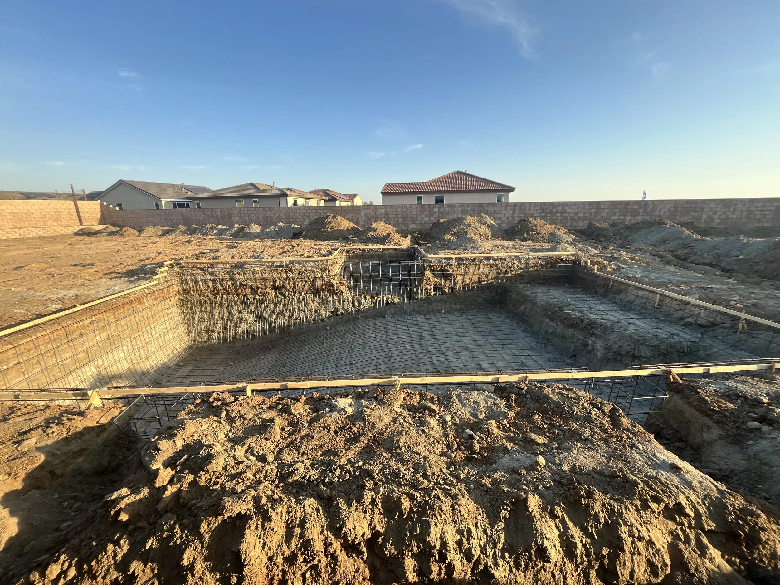 Construction site with foundation work and rebar for a building, surrounded by dirt piles, in a neighborhood with houses in the background, during sunset.