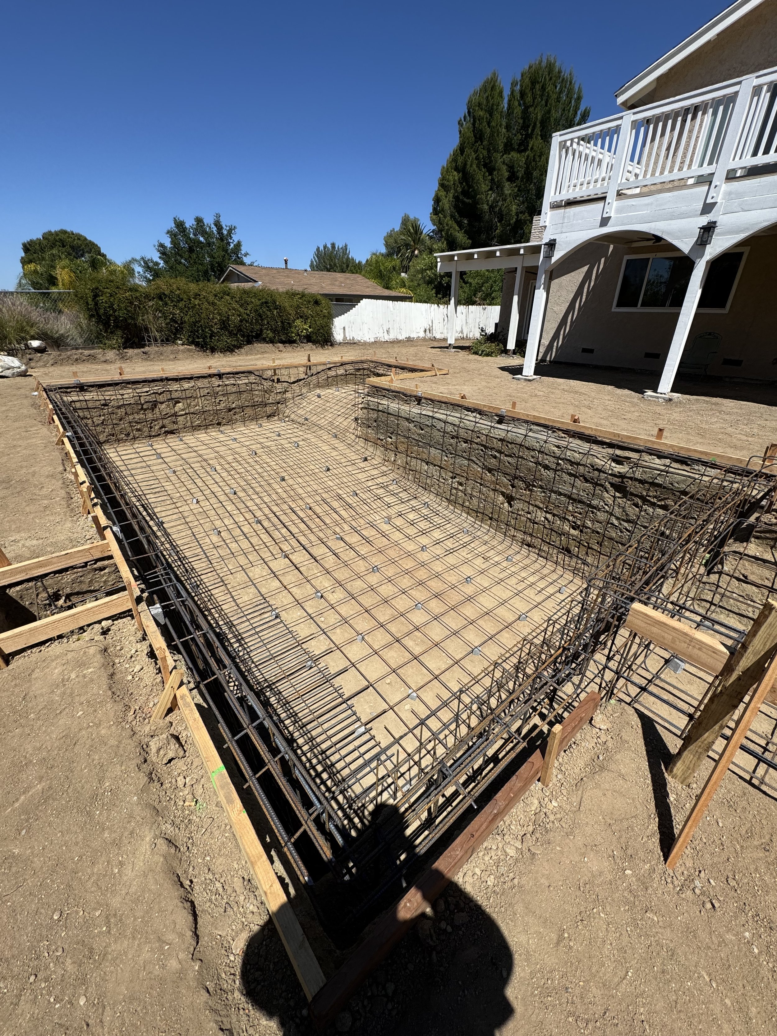 Construction site of a swimming pool with rebar framework and wooden formwork in a backyard.
