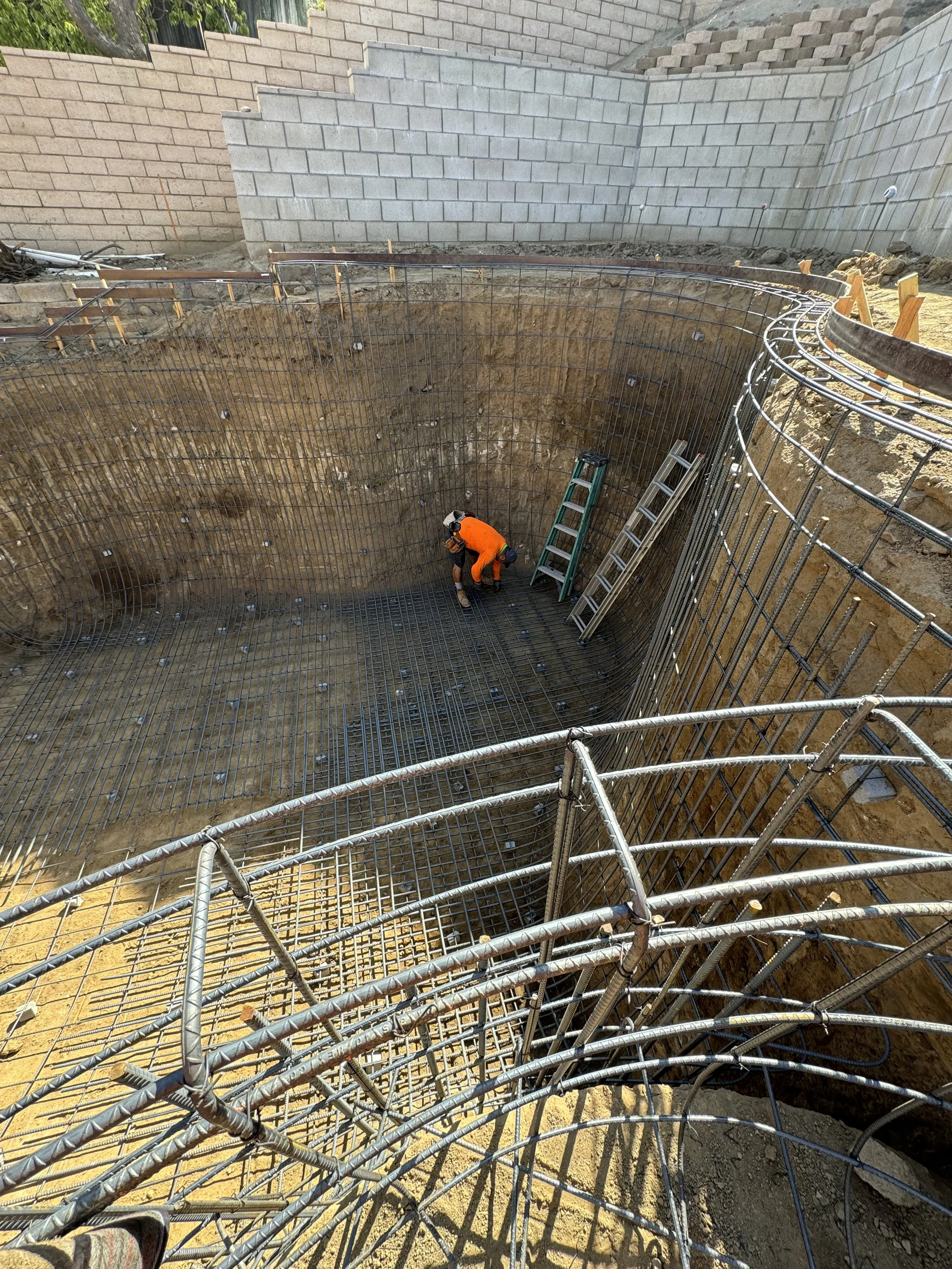 Construction worker in an orange safety vest and helmet working inside a deep excavation site reinforced with steel rebar, with ladders leaning against the wall.
