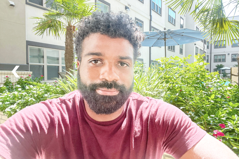 A man with a beard and curly hair taking a selfie outdoors in a courtyard with green plants and palm trees, under a blue umbrella, with apartment buildings in the background.