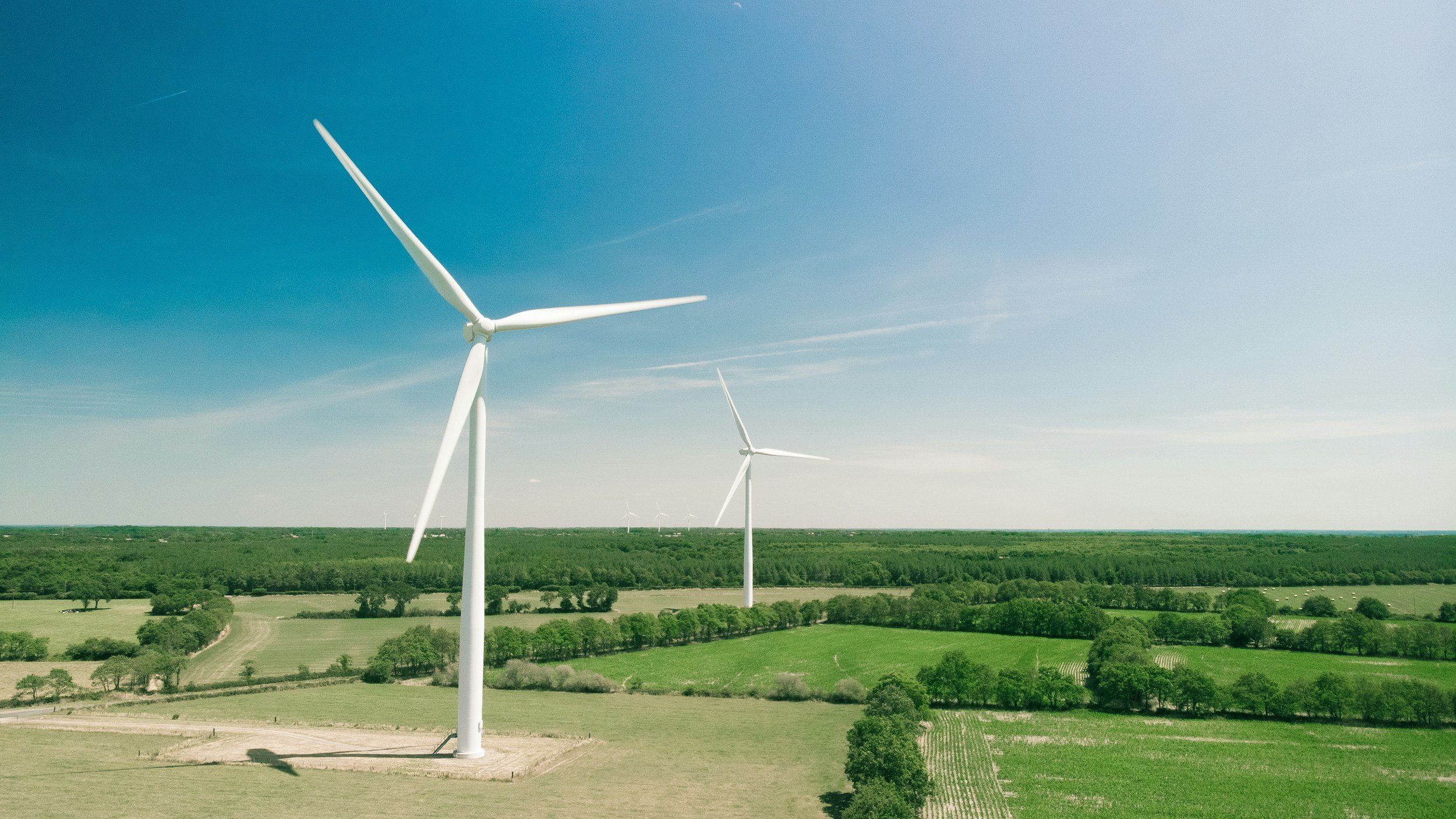 A landscape of green fields and trees under a clear blue sky with large white wind turbines.