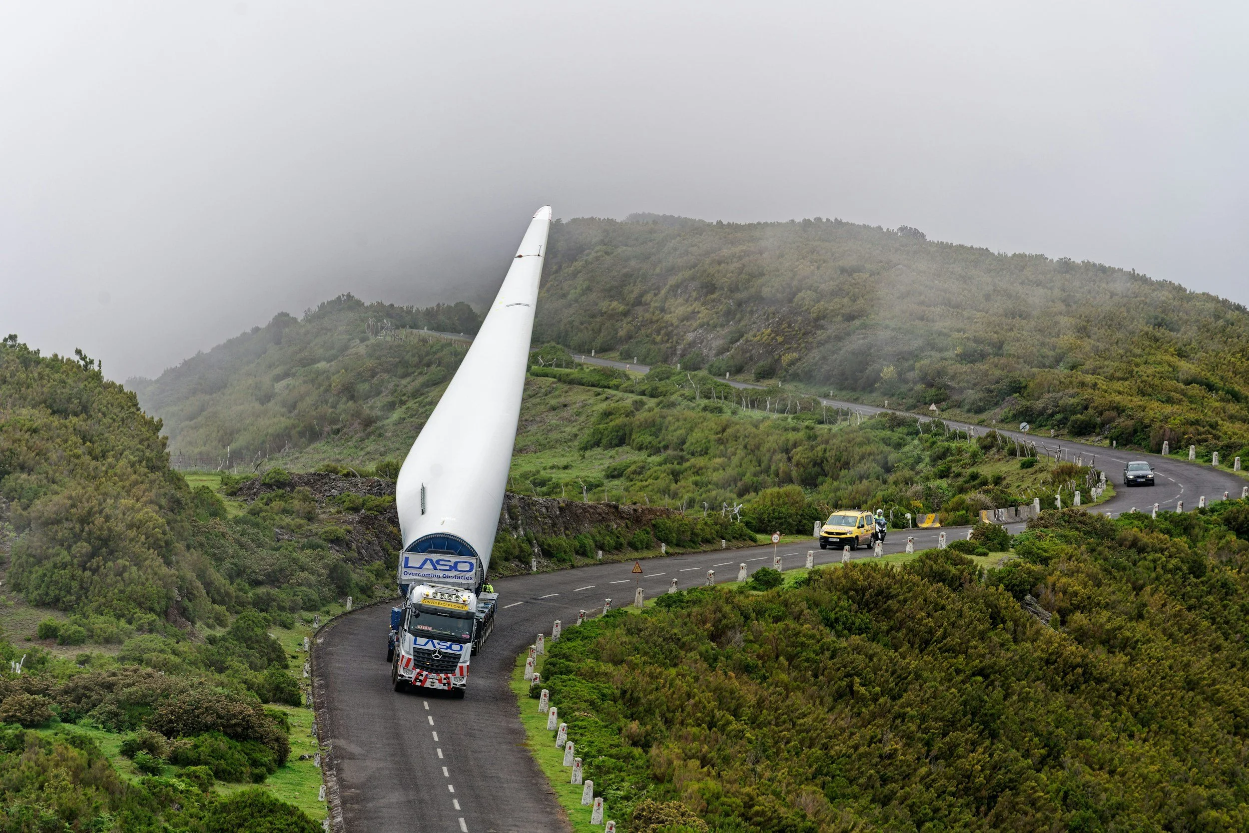 A large truck labeled LASO is transporting a wind turbine blade along a winding mountain road surrounded by green hills and dense vegetation, with foggy weather overhead.