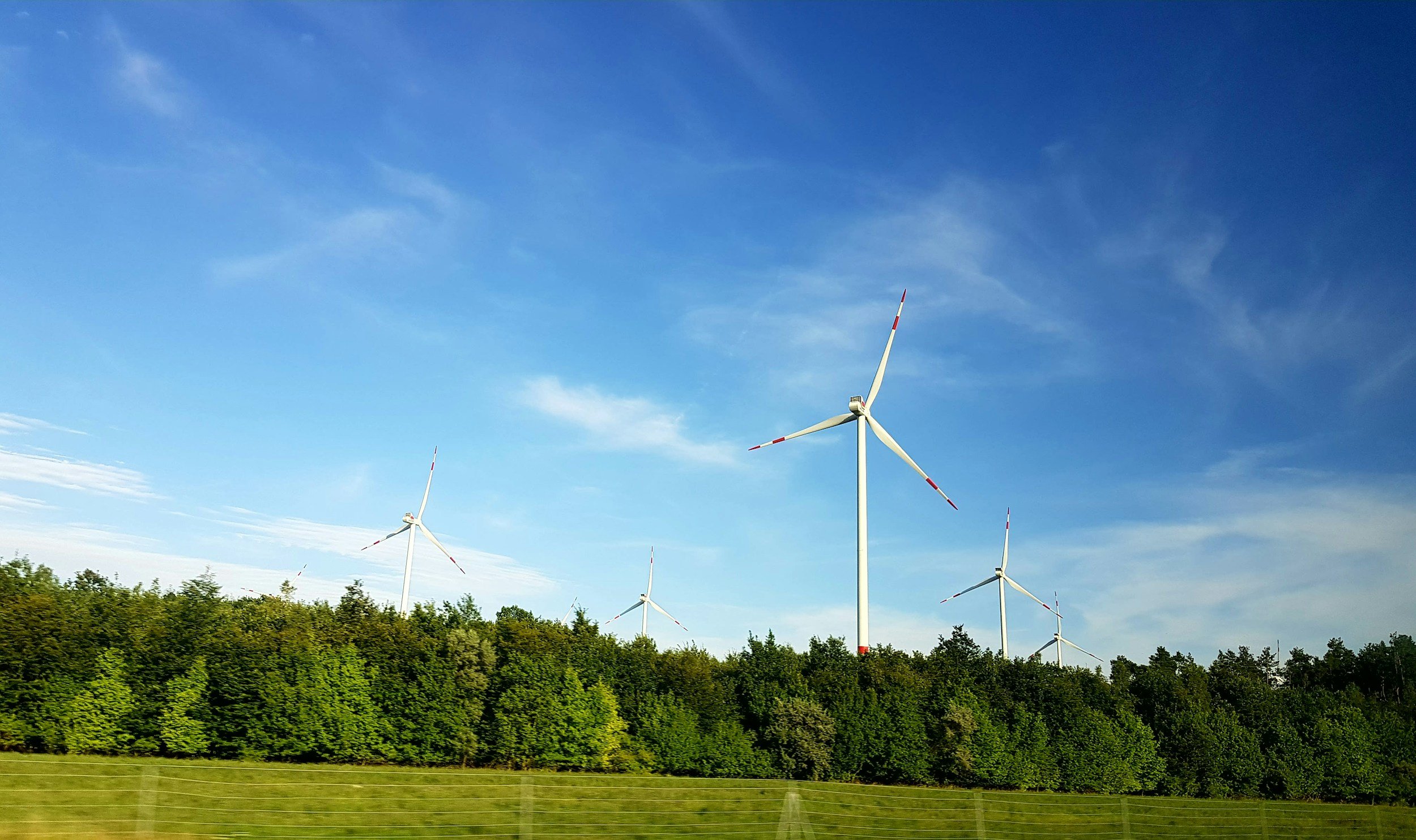 Four wind turbines on a hillside with green trees under a partly cloudy blue sky.