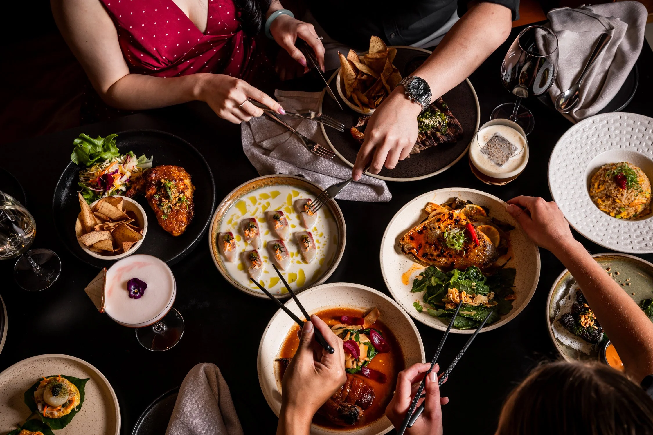 A restaurant table set with various dishes including salads, spring rolls, tempura, and a cocktail, against a wall with a black-and-white mural of a street scene.