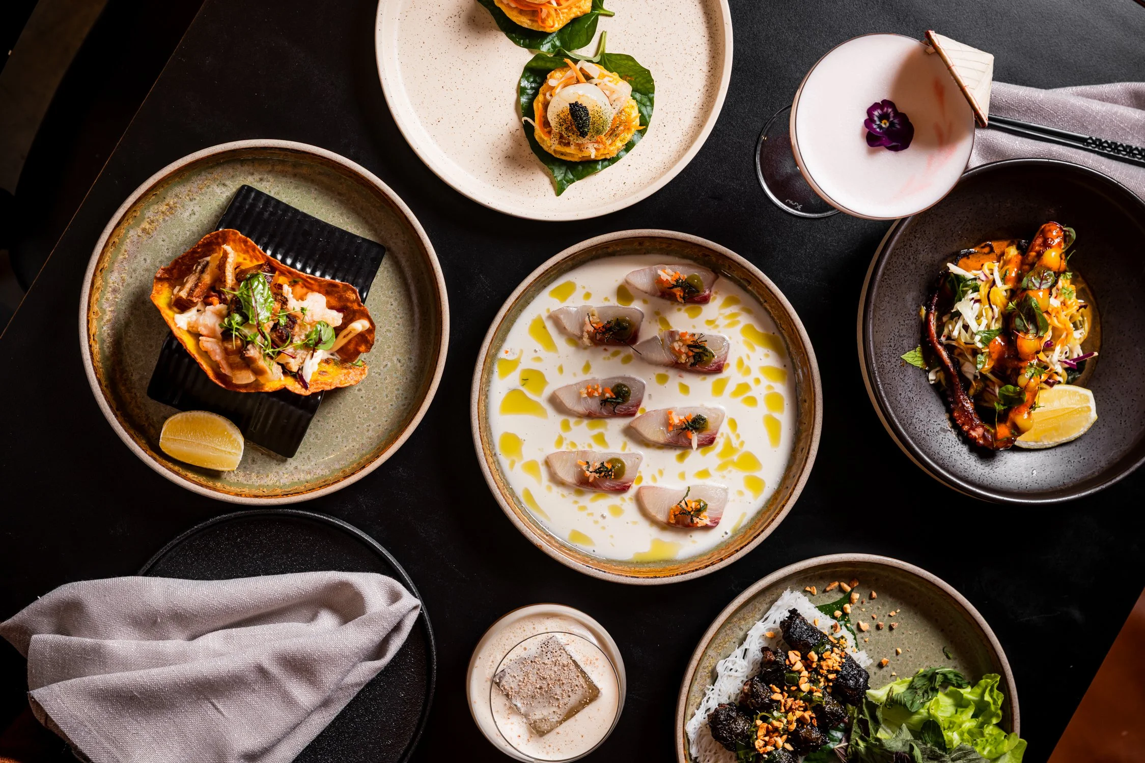 A restaurant table set with various dishes including salads, spring rolls, tempura, and a cocktail, against a wall with a black-and-white mural of a street scene.