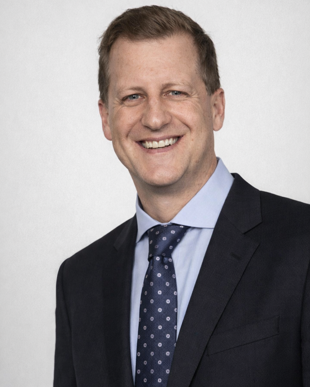 A smiling man with short brown hair, wearing a dark suit, light blue shirt, and navy tie with white polka dots.