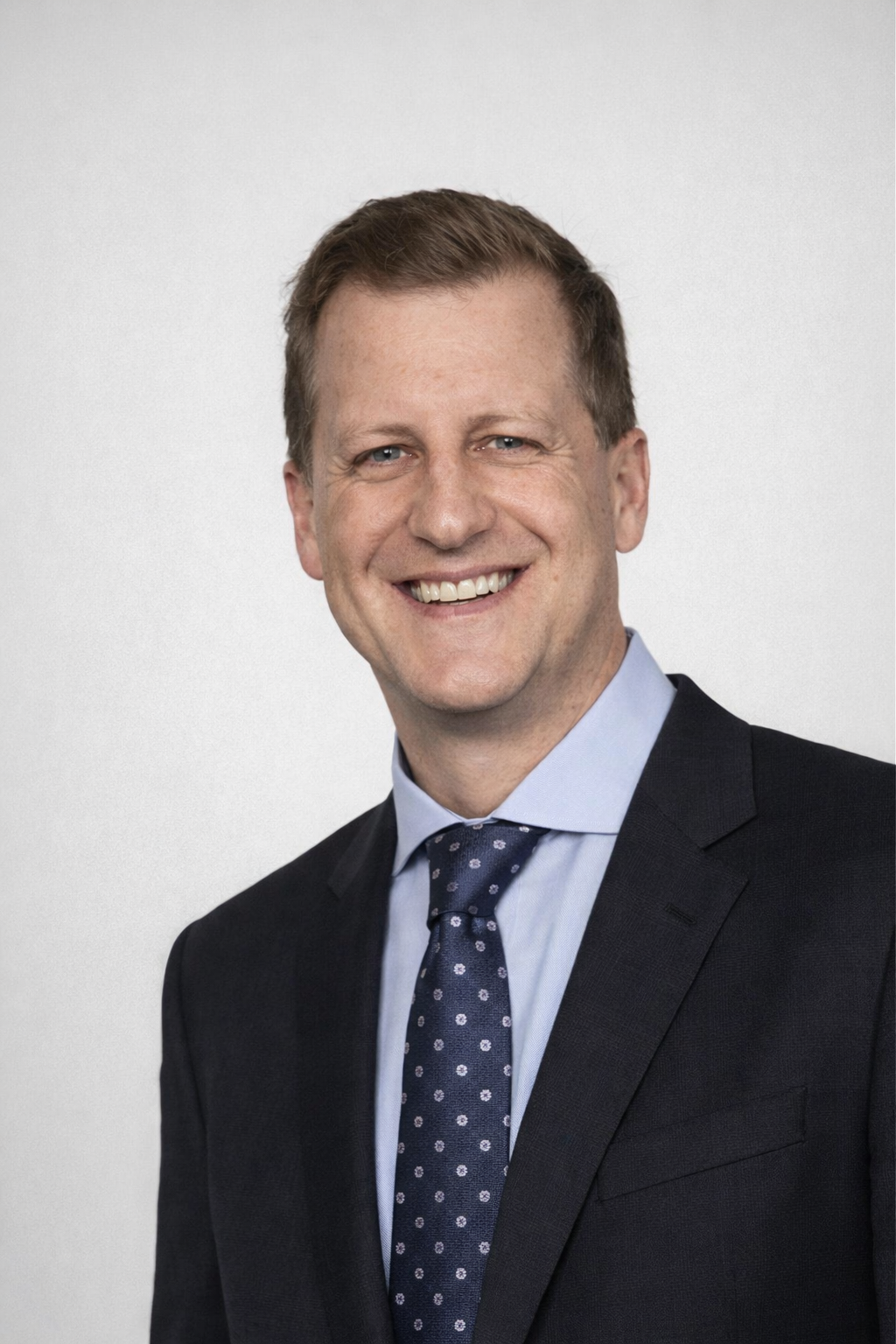 A smiling man with short brown hair, wearing a dark suit, light blue shirt, and navy tie with white polka dots.