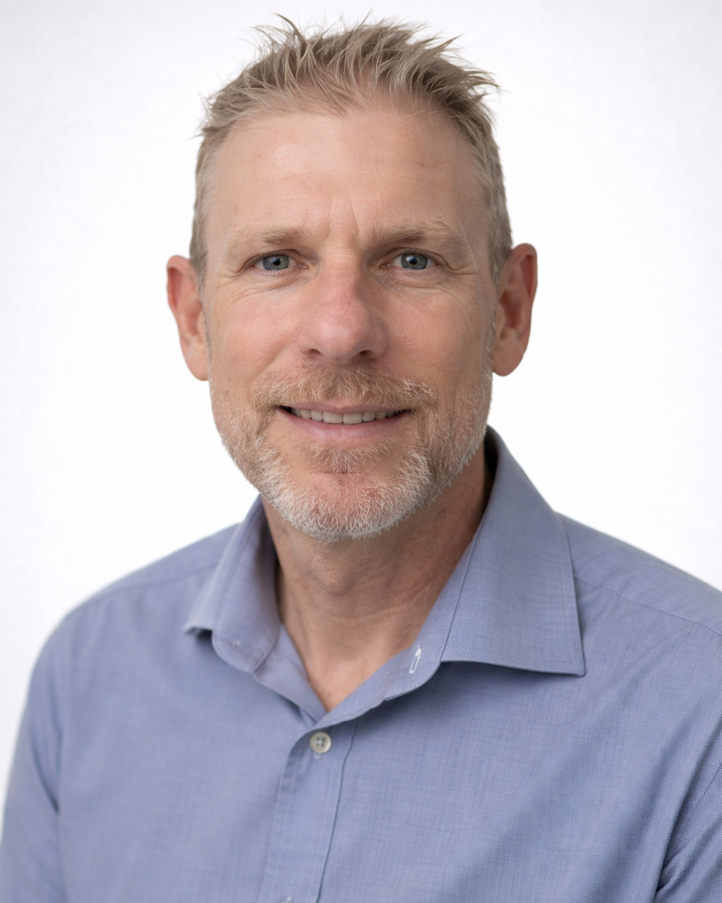A man with short brown hair wearing a white and blue checkered button-up shirt standing against a plain beige wall.