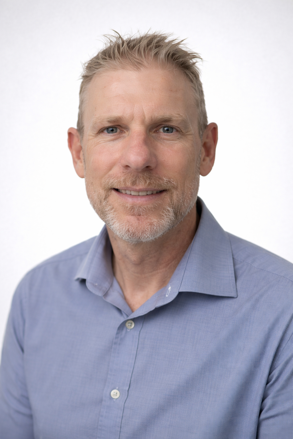 A man with short brown hair wearing a white and blue checkered button-up shirt standing against a plain beige wall.