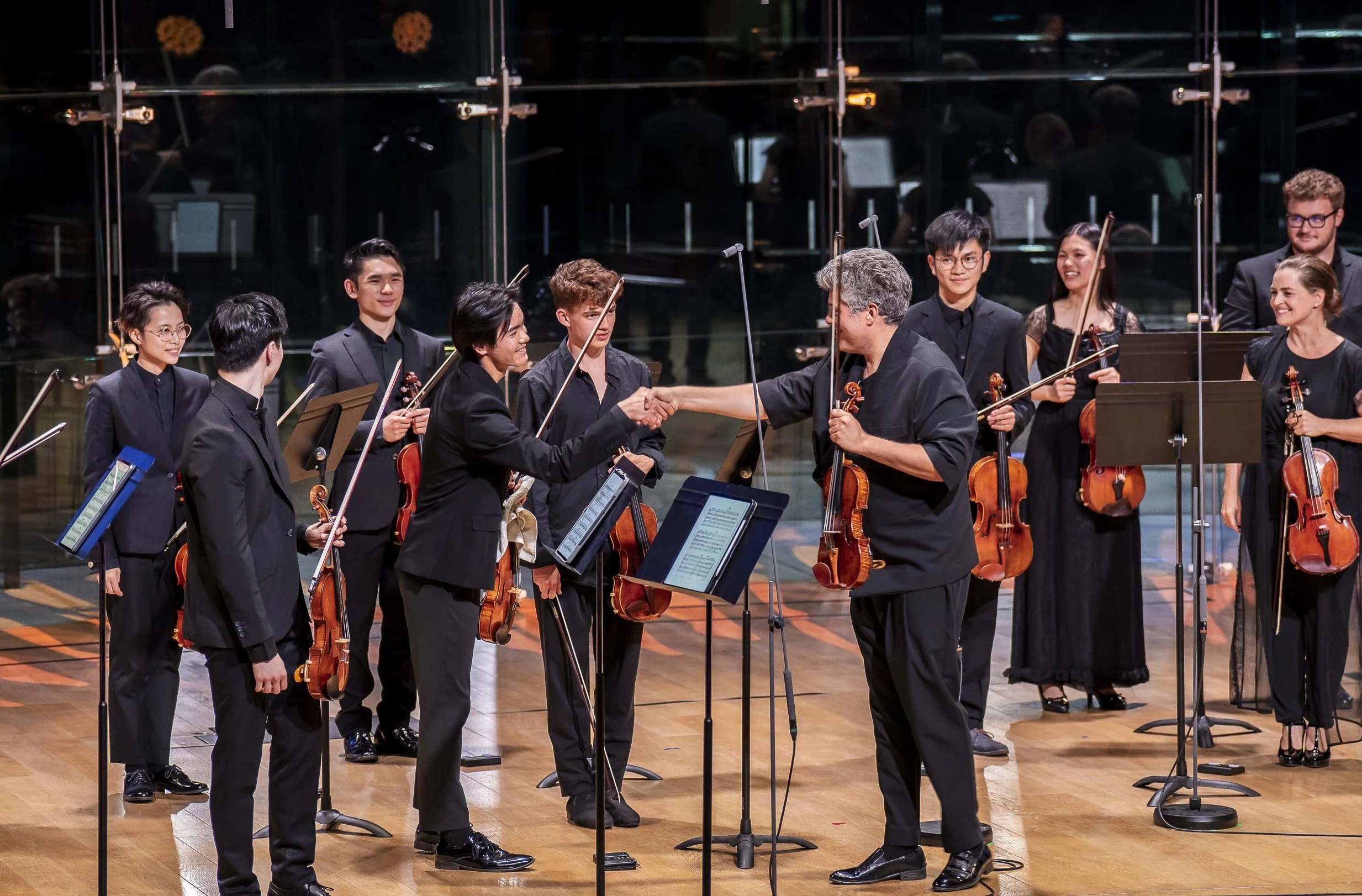 A group of musicians, mostly young adults, on stage with their violins, accepting a handshake from an older conductor after a performance, with music stands and a reflective black background.