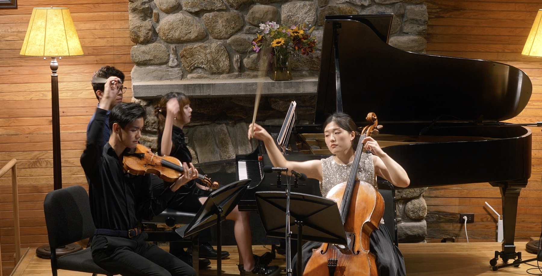 Three young women and one young man playing classical music instruments in an indoor setting with wooden walls, a fireplace, and a grand piano. One woman plays the cello, another violin, and a third violin, while a man plays the violin.