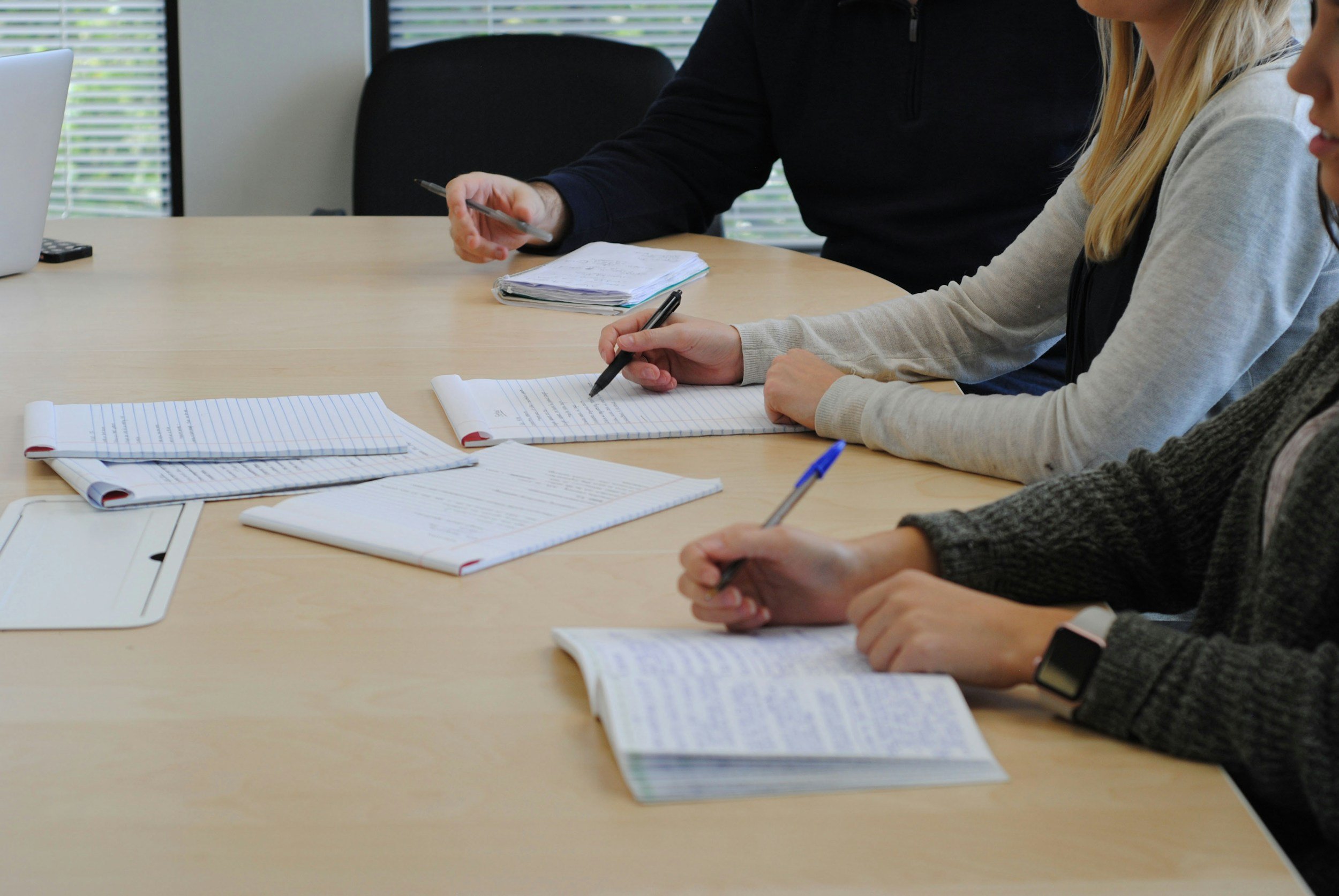 Meeting with three people sitting at a table with notebooks and pens, discussing or taking notes.