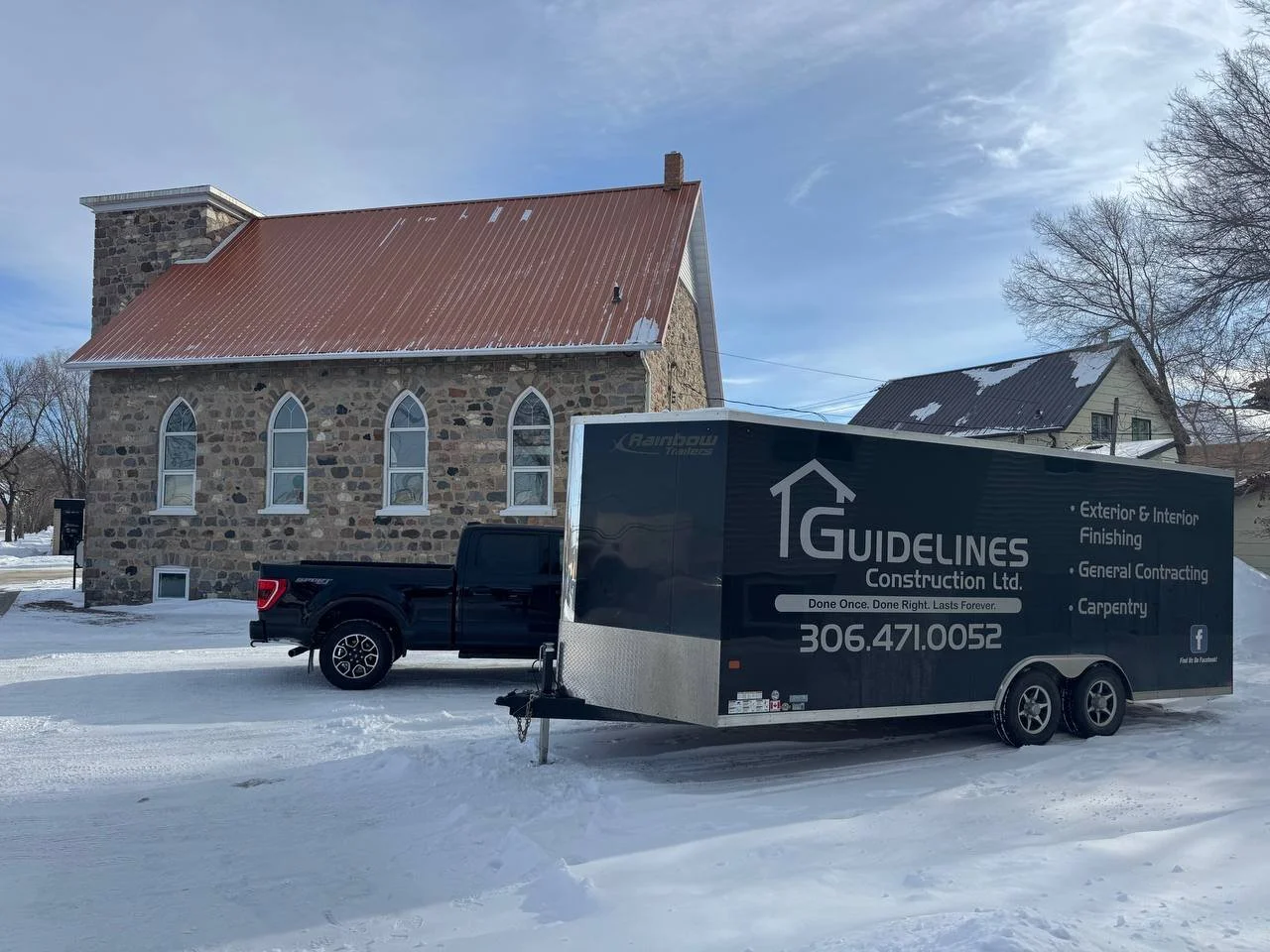 A black pickup truck with a large gray trailer attached, parked on a snowy street in front of a stone church with a red metal roof and arched windows on a clear winter day.