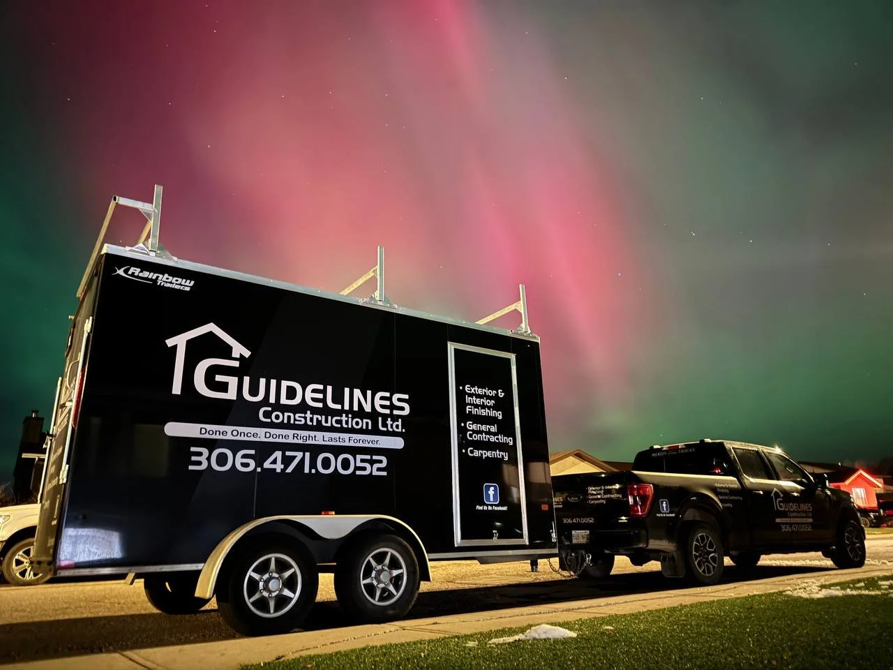 Night scene with a truck and trailer parked on the street, advertising Guidelines Construction with the Aurora Borealis visible in the sky.