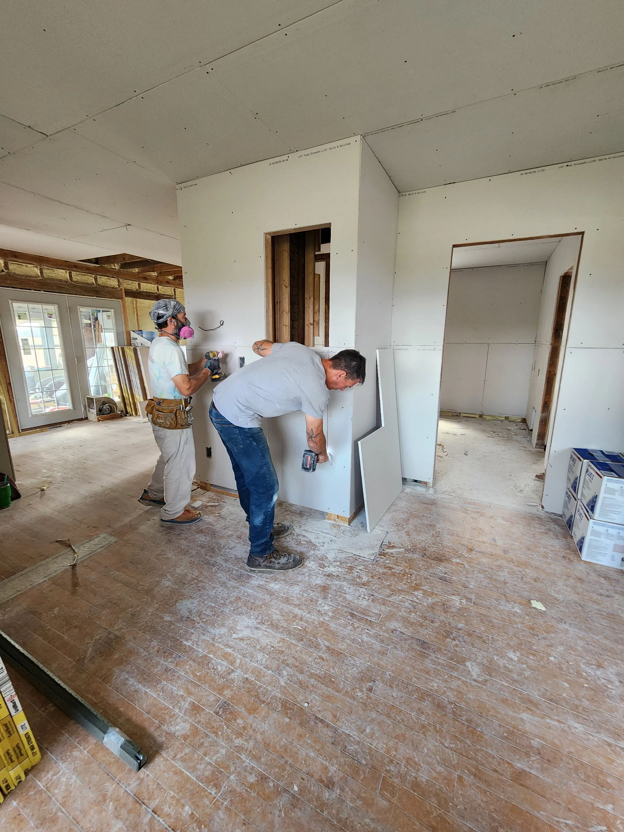 Two construction workers installing drywall in an unfinished room of a house under renovation.