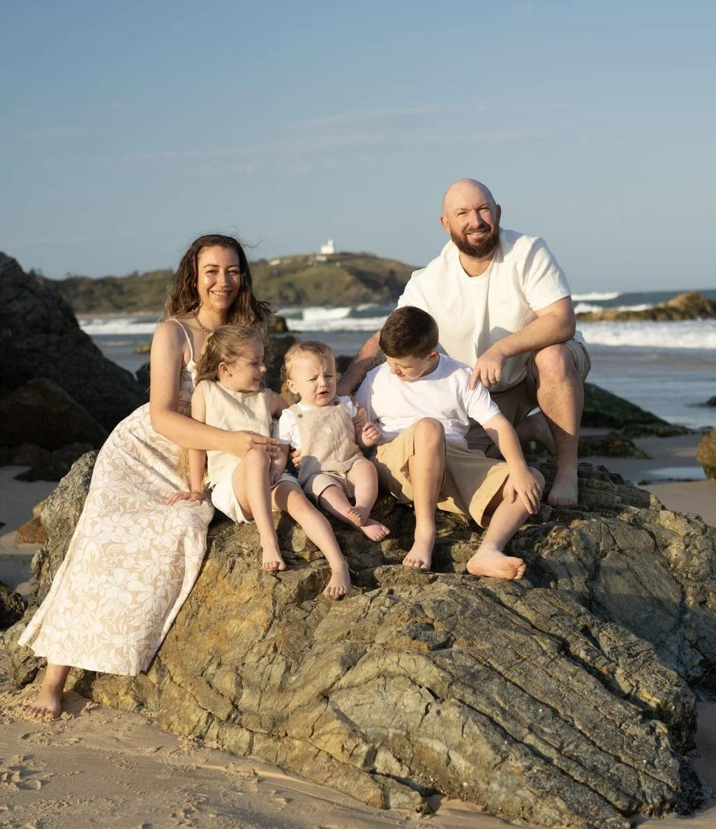 A family of six sitting on rocks at the beach during sunset, with ocean waves in the background, smiling and enjoying their time together.