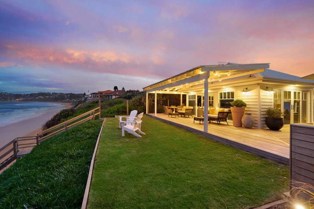 View of a house with a deck overlooking a beach at sunset, with outdoor furniture and large potted plants on the deck.