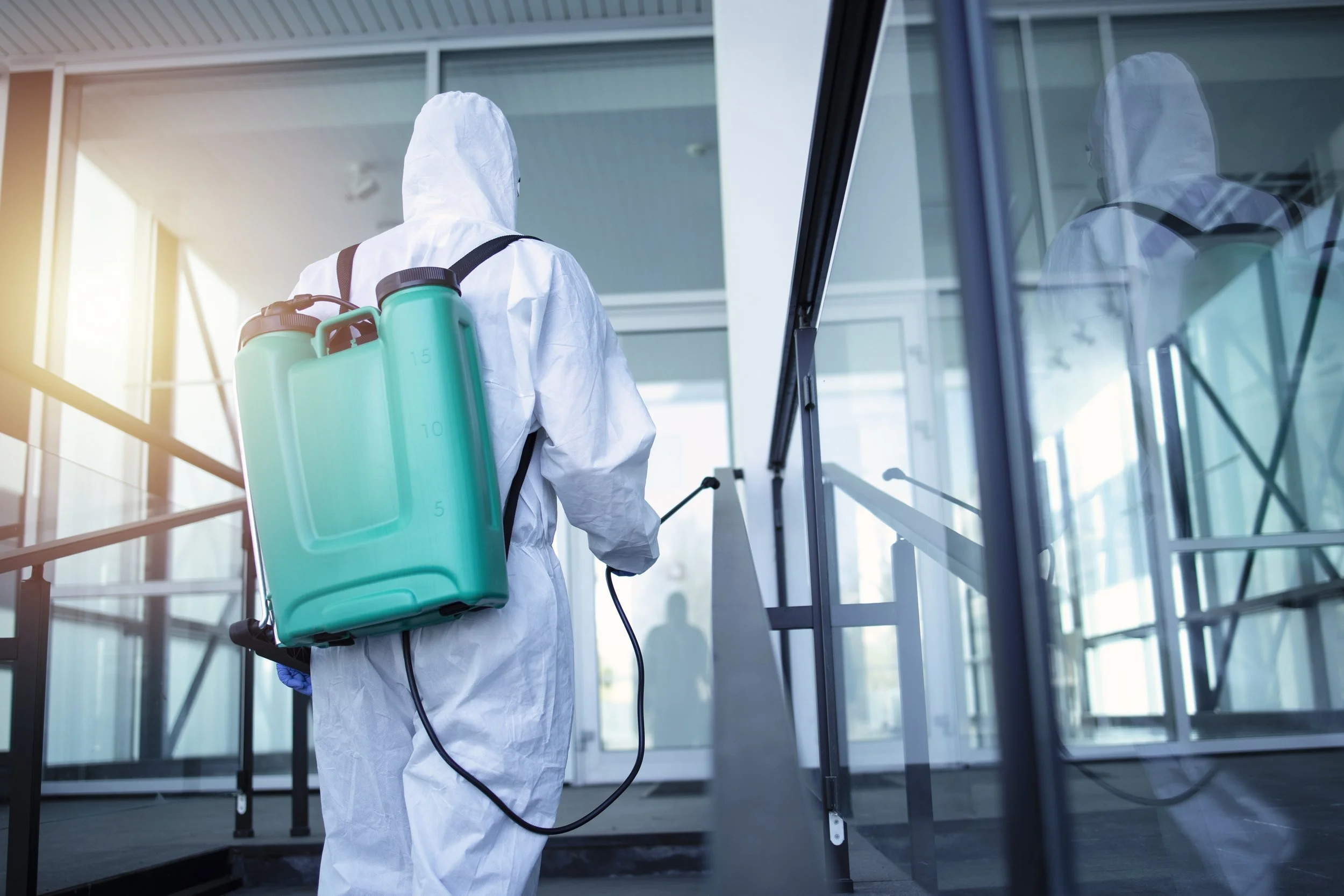 A person wearing a white protective suit and a backpack sprayer disinfects the exterior of a building.
