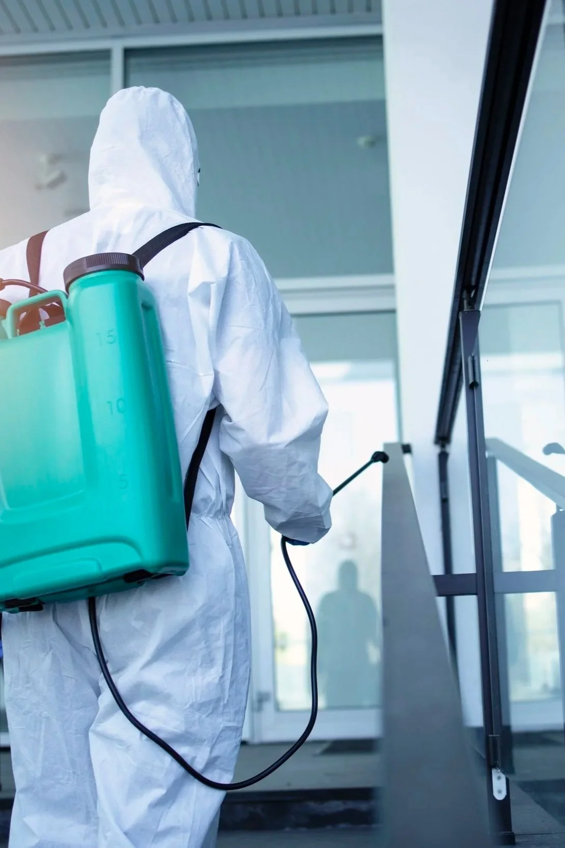 Person in full protective gear disinfecting a building entrance with a spray wand.
