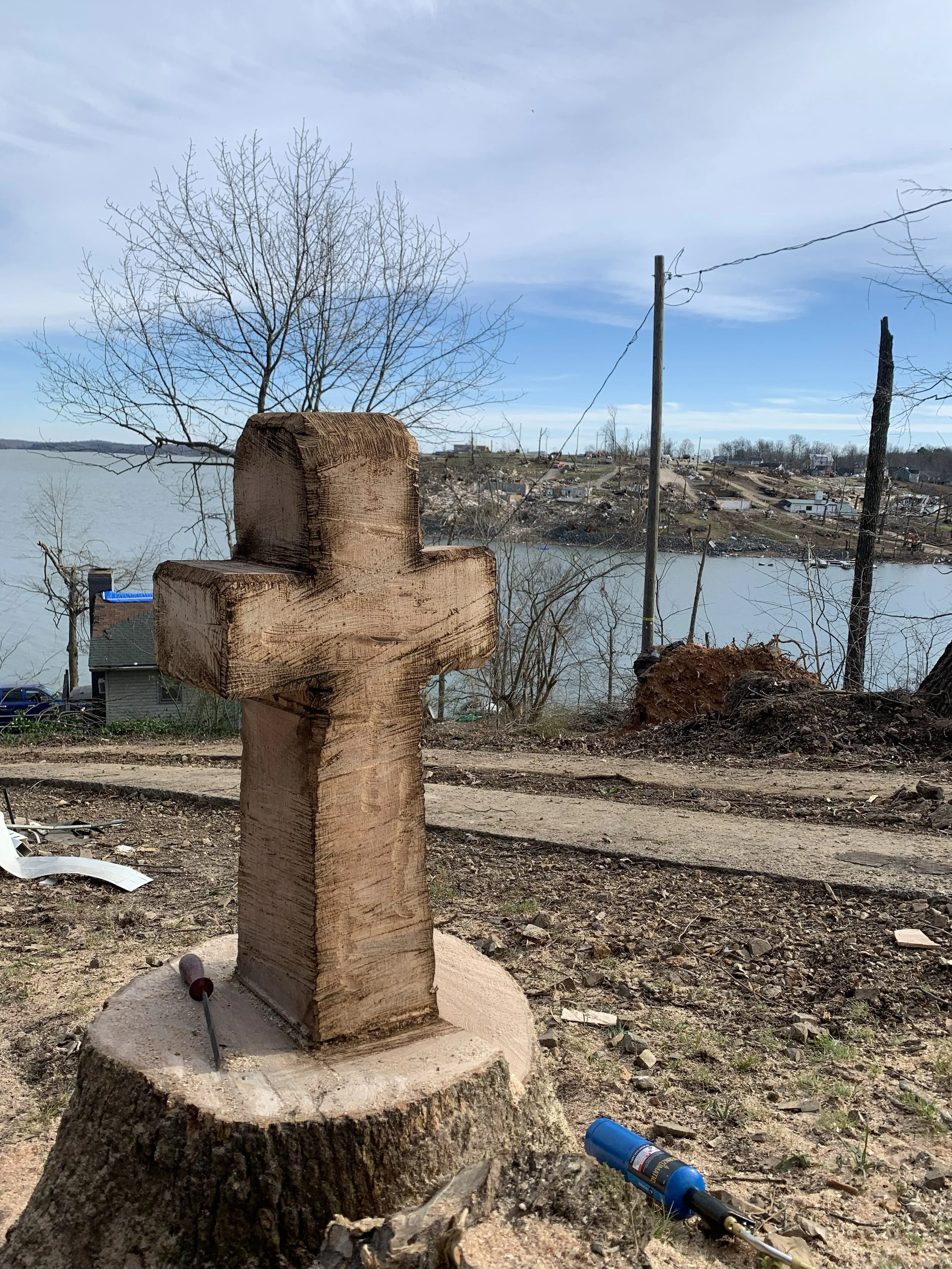 A wooden cross carved from a tree trunk stands on a tree stump with a screwdriver and a power drill on the ground nearby. In the background, there is a river, leafless trees, and a hilly area with houses and power lines under a partly cloudy sky.