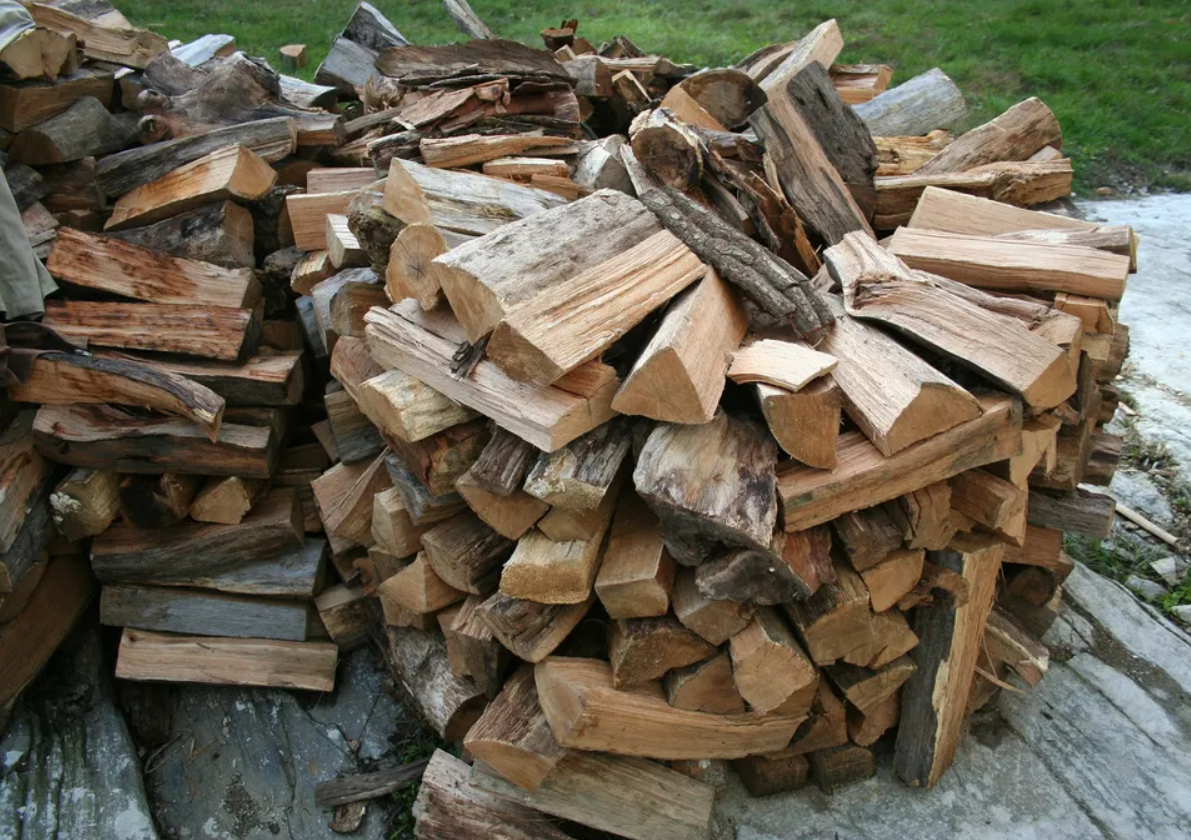 A large pile of split and chopped firewood stacked outdoors on a paved surface with grass in the background.