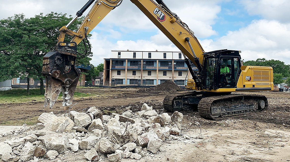 A yellow Caterpillar excavator with a hydraulic hammer attachment breaking rocks on a construction site. There is a pile of rubble in front of the excavator and an apartment building in the background under a cloudy sky.