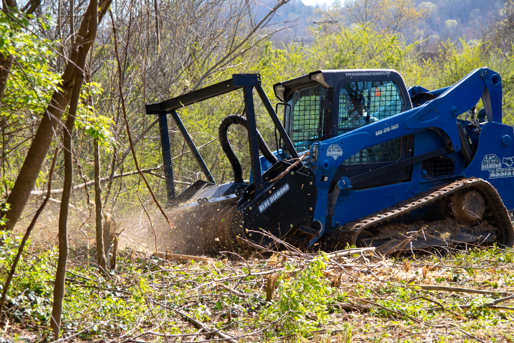 Blue tracked skid steer loader clearing overgrown vegetation in a wooded area.