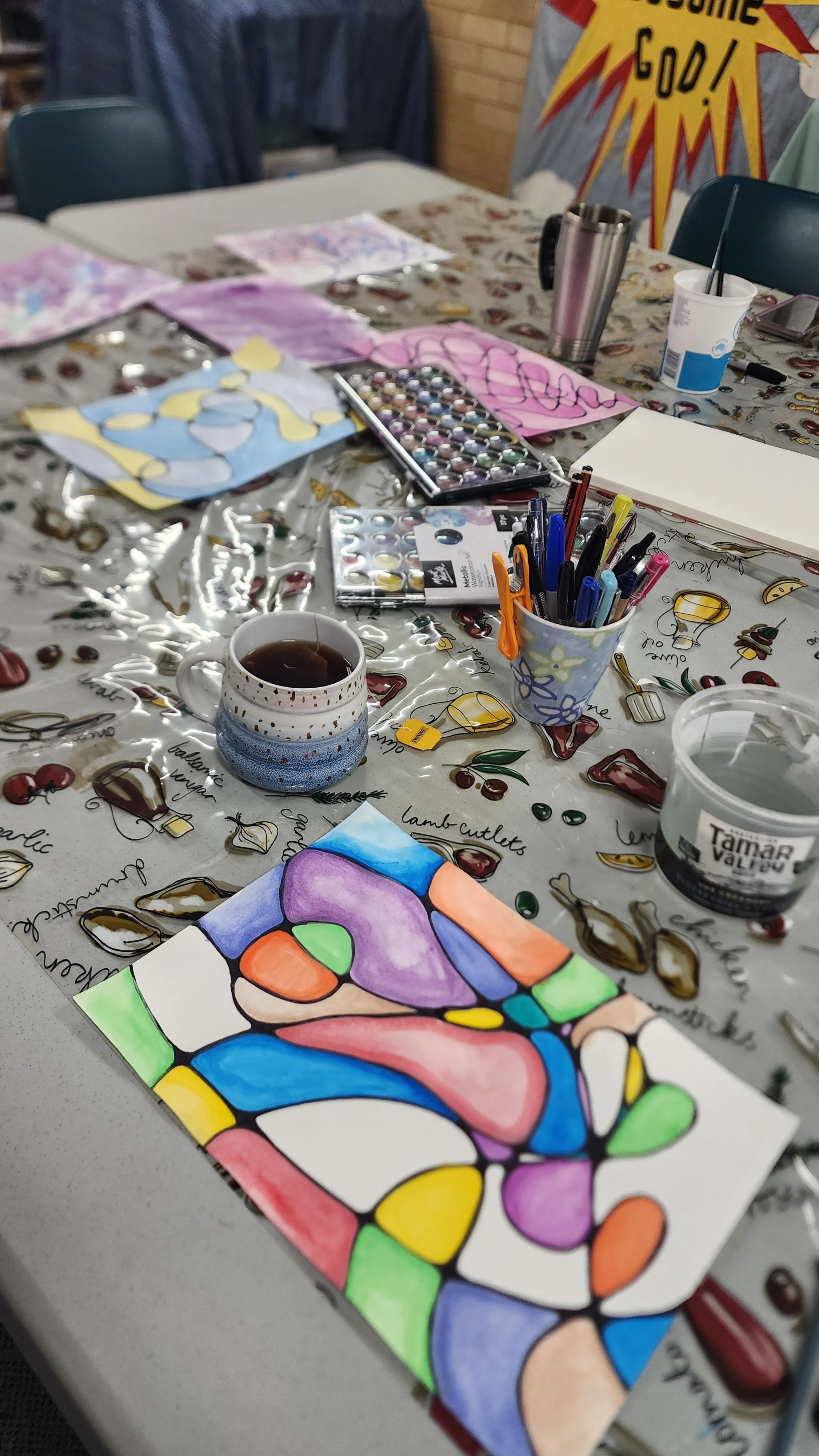 Colorful abstract watercolor painting on paper, scattered art supplies including pens, watercolor paints, paper, coffee cup, and various art projects on a table covered with cherry-patterned tablecloth in a craft room.