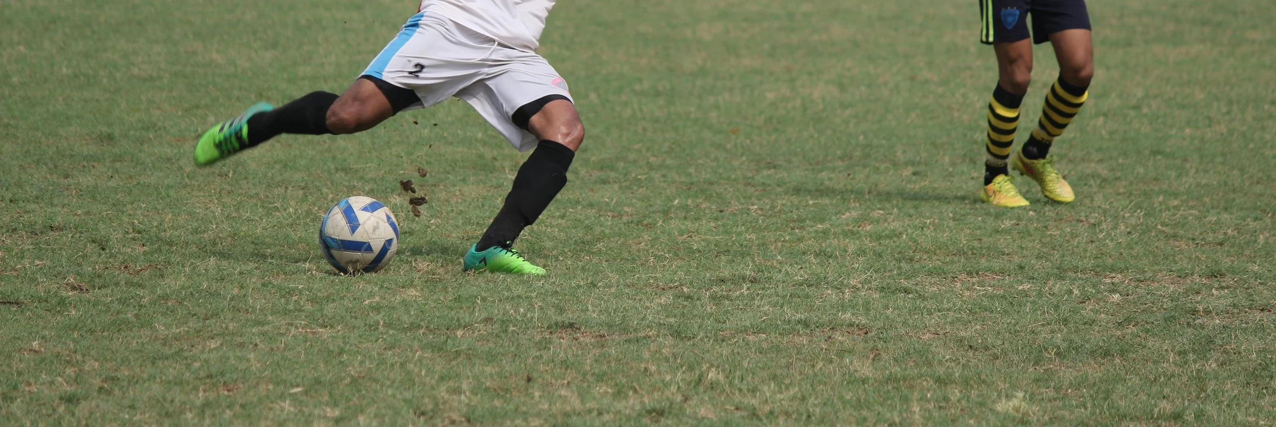 Soccer player kicking a ball on the field while another player approaches, only their legs visible.