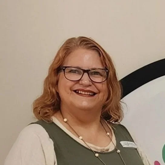 A photo of Caroline Hochstetter - a woman with shoulder-length red hair and glasses smiling at the camera, wearing a white shirt with a green vest and a name tag.