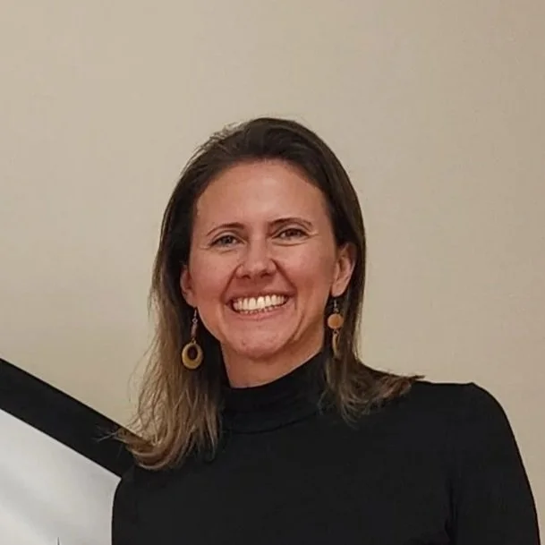 A photo of Angela Ngiam - a woman with shoulder-length brown hair, smiling, wearing a black turtleneck and gold hoop earrings.