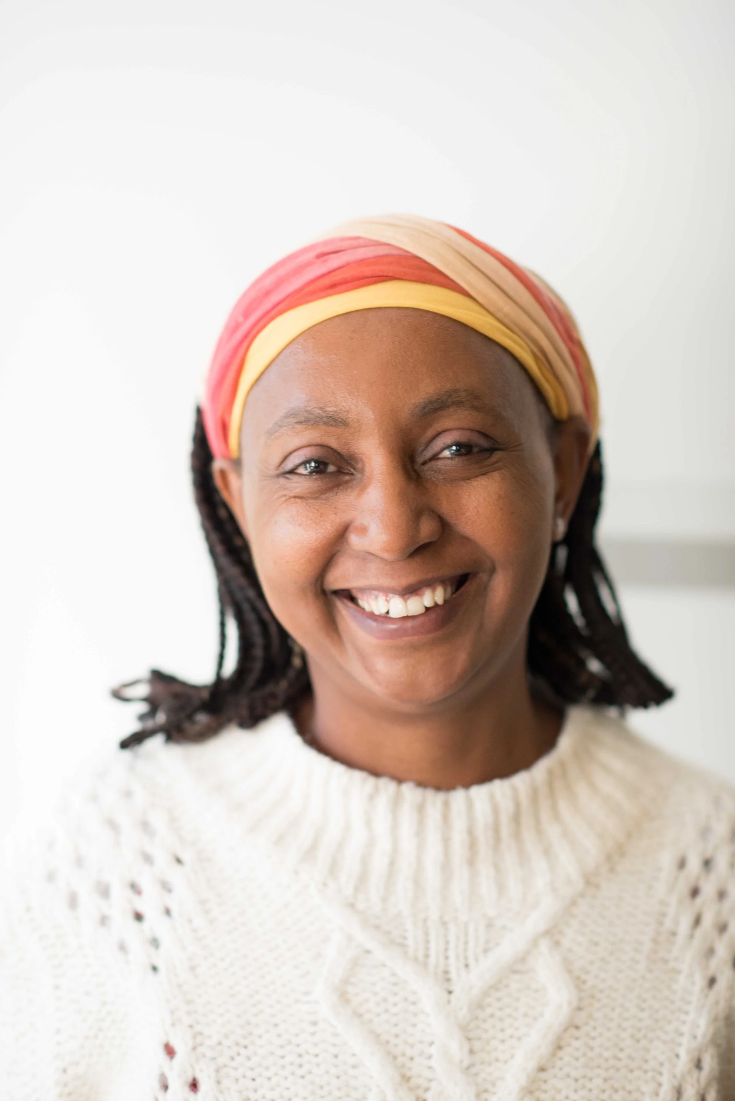 A photo of Lydia Gitau - a smiling woman wearing a colorful headscarf and a white knitted sweater.