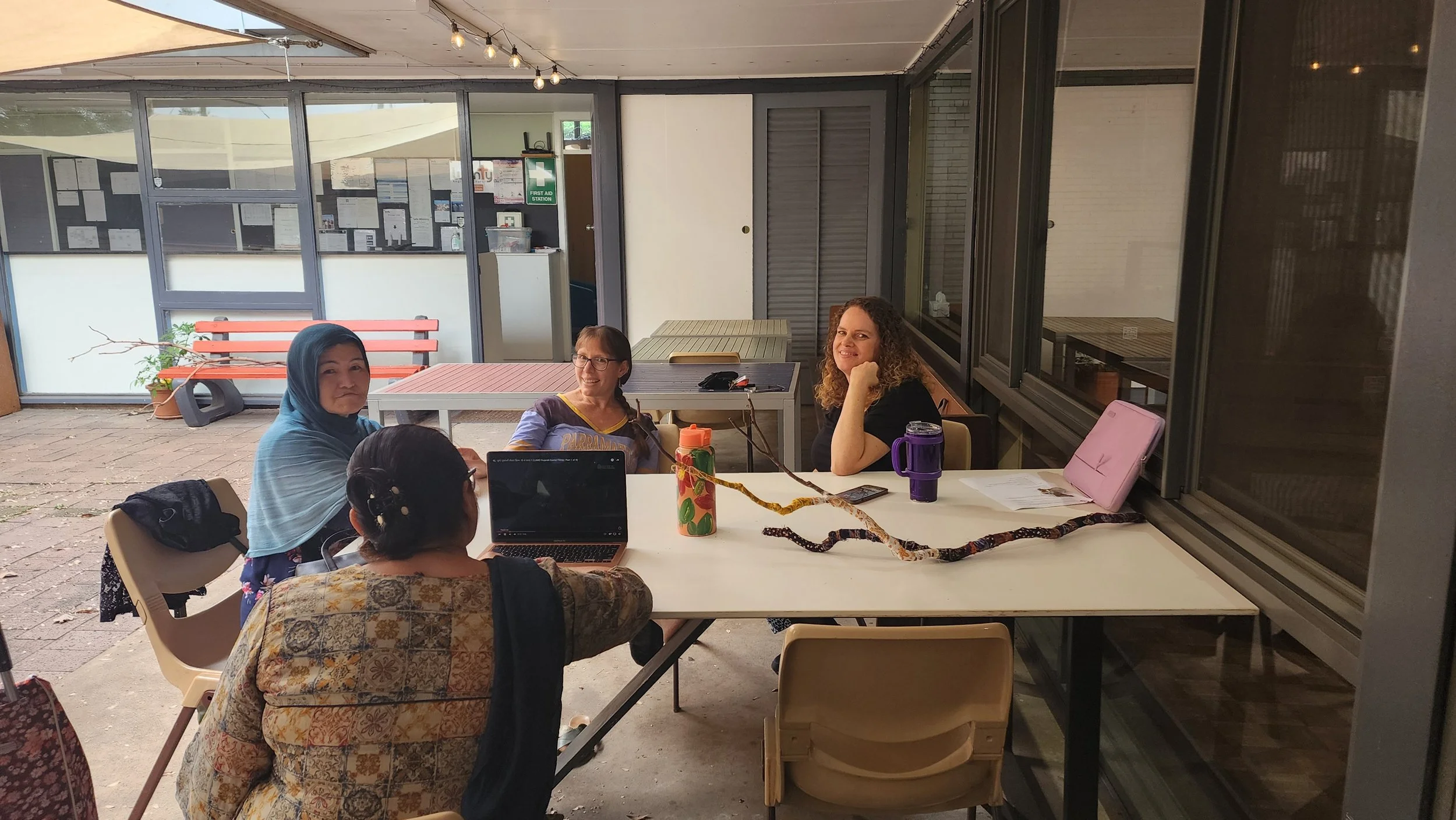 Four women sitting around a table in an indoor-outdoor space, engaged in a conversation or meeting. One woman is wearing a blue headscarf, another has glasses, and there are laptops, water bottles, and papers on the table.