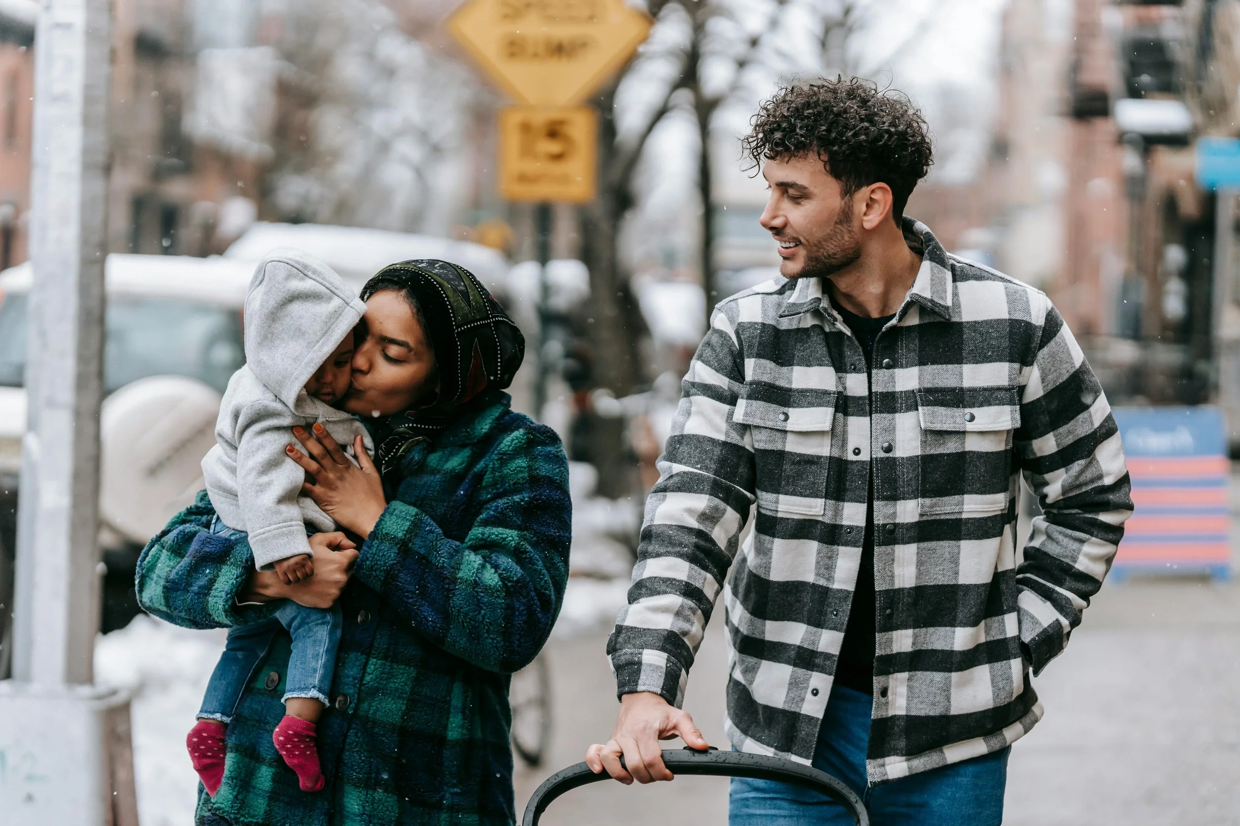 A woman is holding a child and kissing them on the forehead while a man stands next to them, pushing a stroller and smiling at them on a winter day.