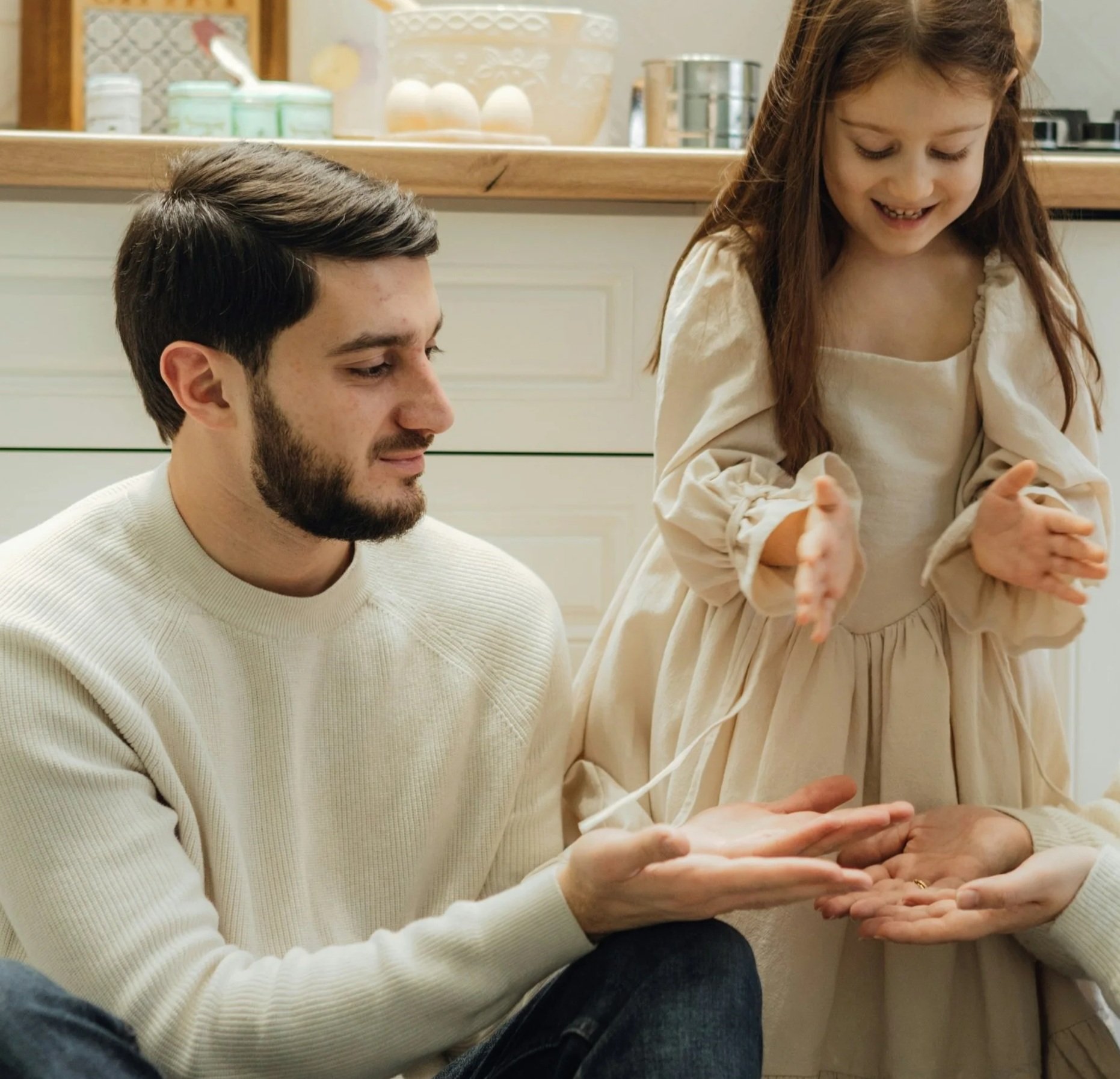A young girl claps her hands while an adult man holding an open hand extends his hand towards her in a warm, indoor setting.
