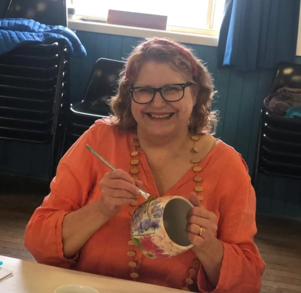 Smiling woman with glasses and curly hair painting a floral design inside a ceramic mug with a paintbrush, seated at a table.