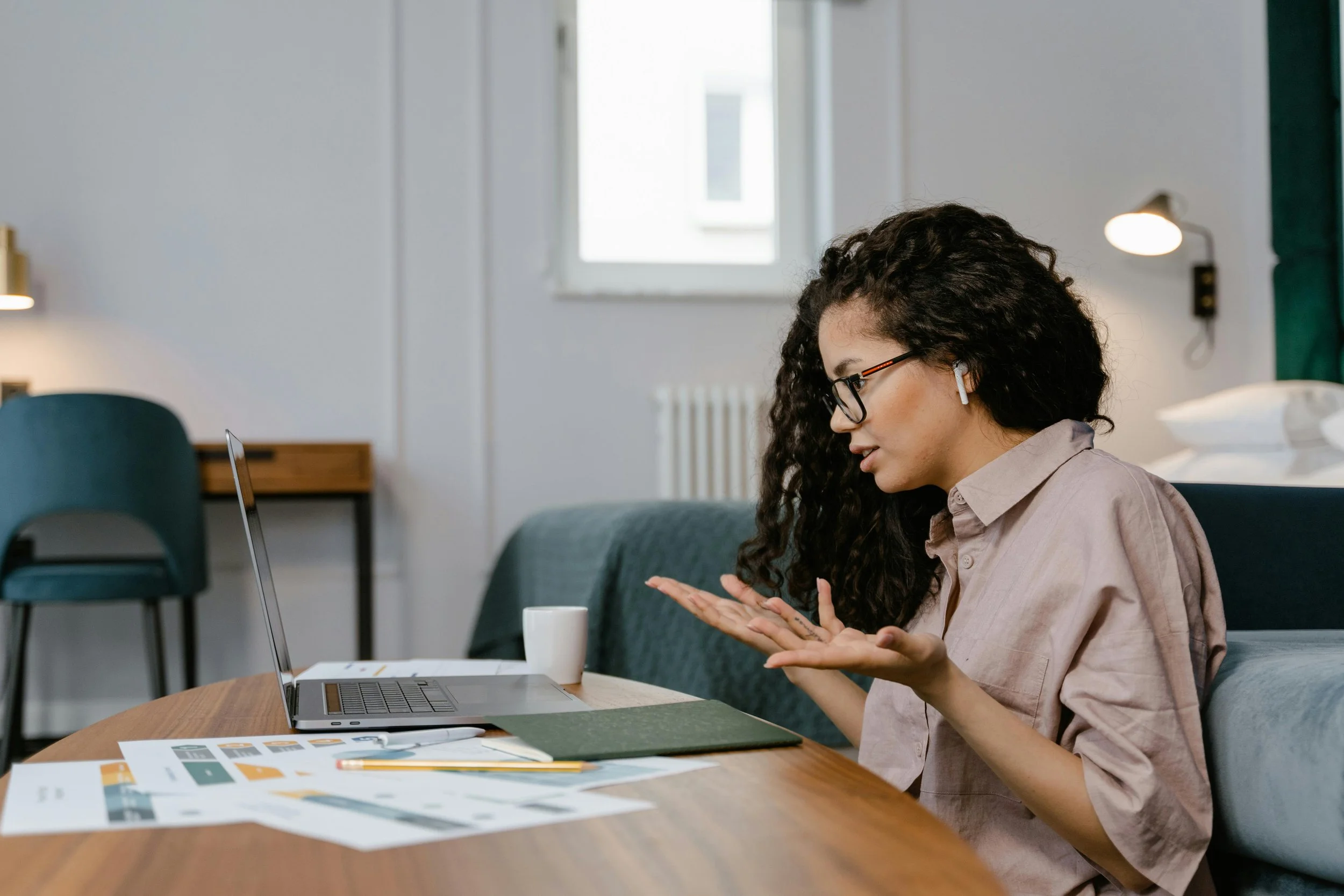 A woman with curly black hair and glasses, sitting at a wooden table in a bright room, working on her laptop. Various papers and a pen are on the table.