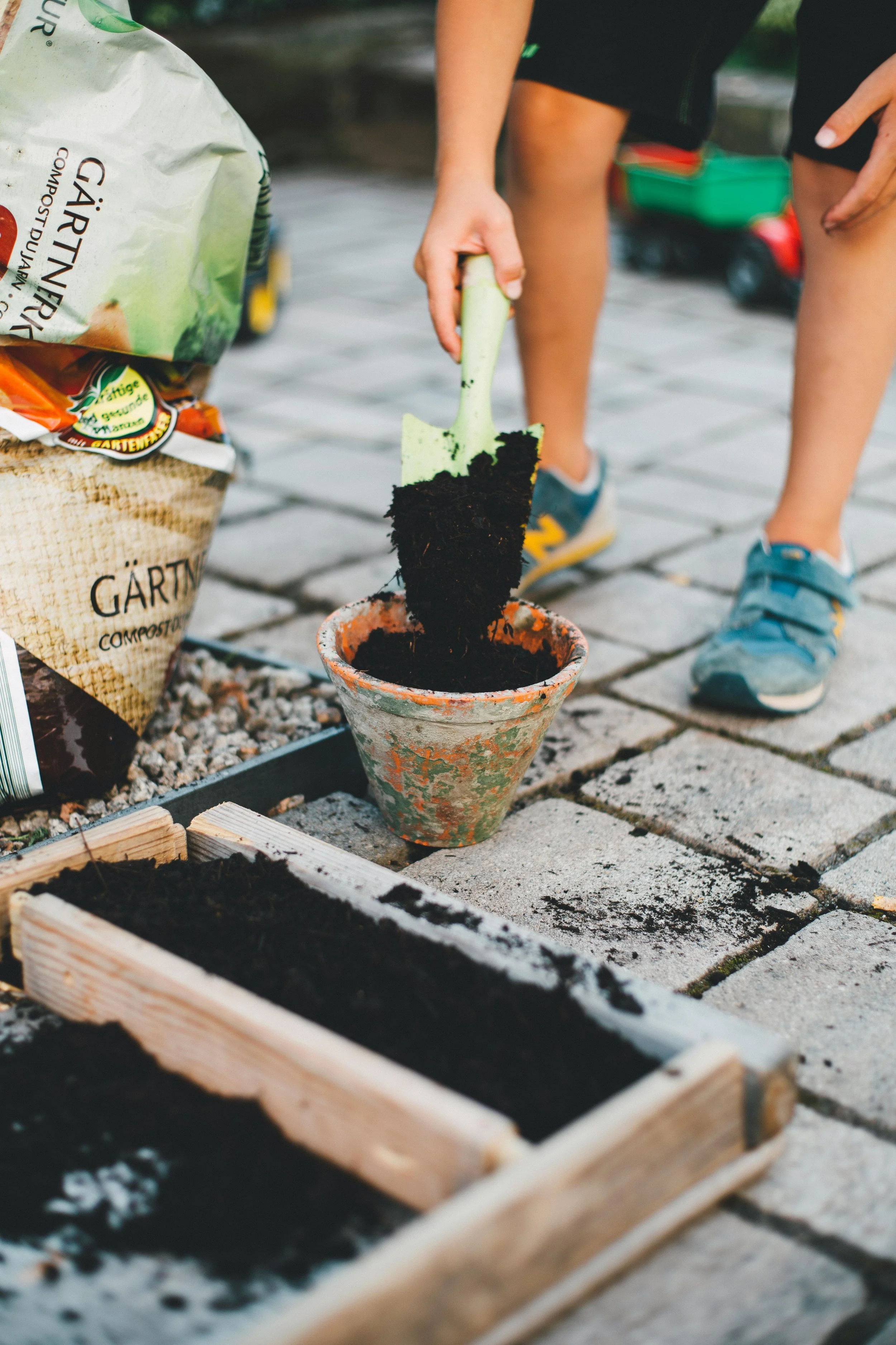 Child planting a small plant into a terracotta pot on a paved outdoor surface, with gardening supplies nearby.