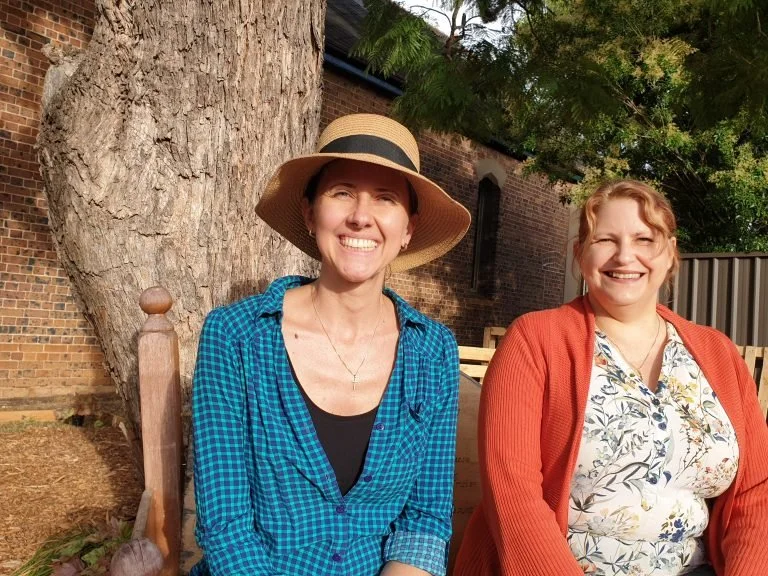 Two women sitting outdoors near a large tree, smiling at the camera. One is wearing a wide-brimmed hat and a blue checked shirt, the other is wearing a floral blouse and an orange cardigan.