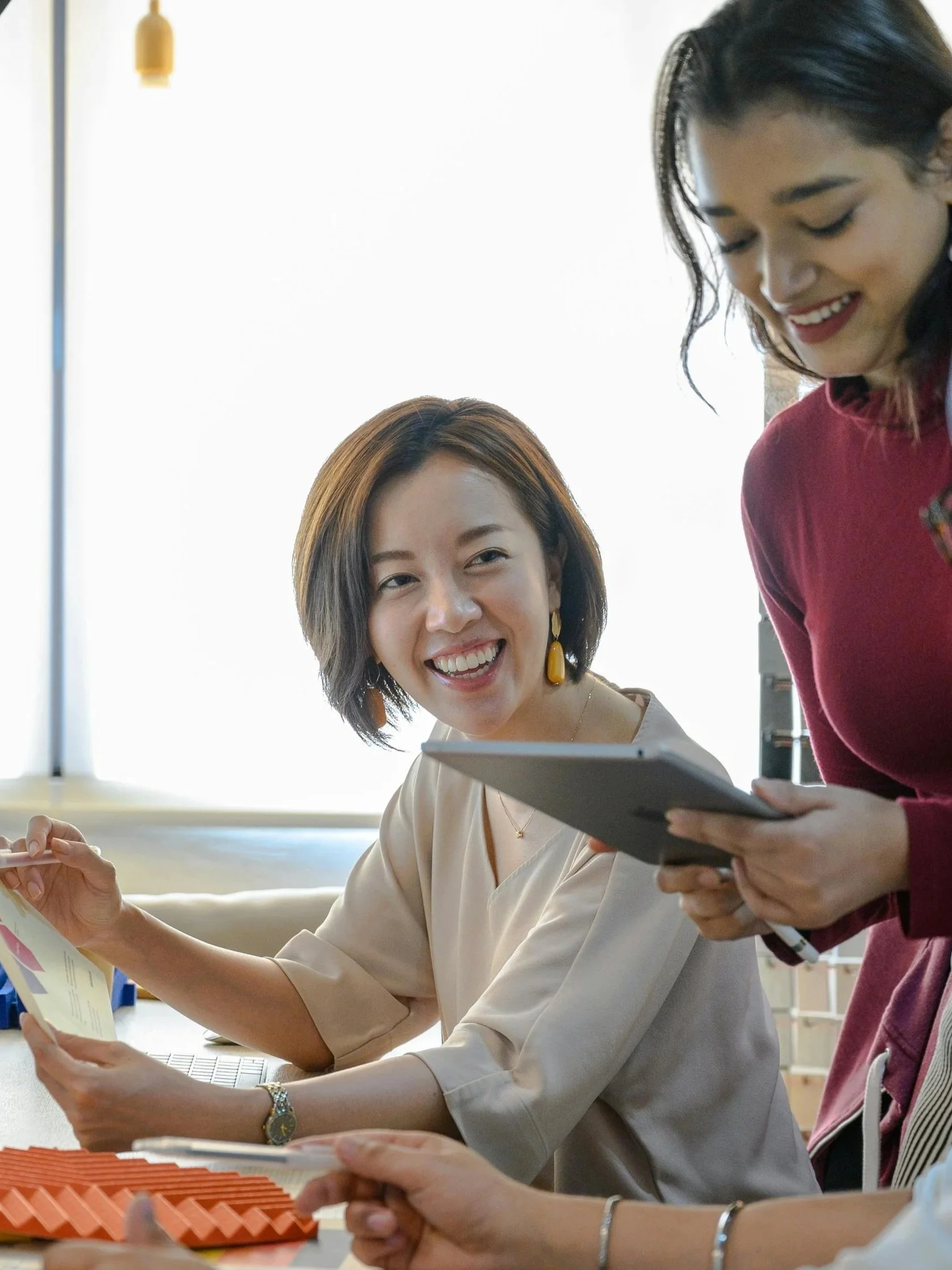Two women engaging in a conversation, one woman smiling and holding a document, while the other holds a tablet in a bright office setting.