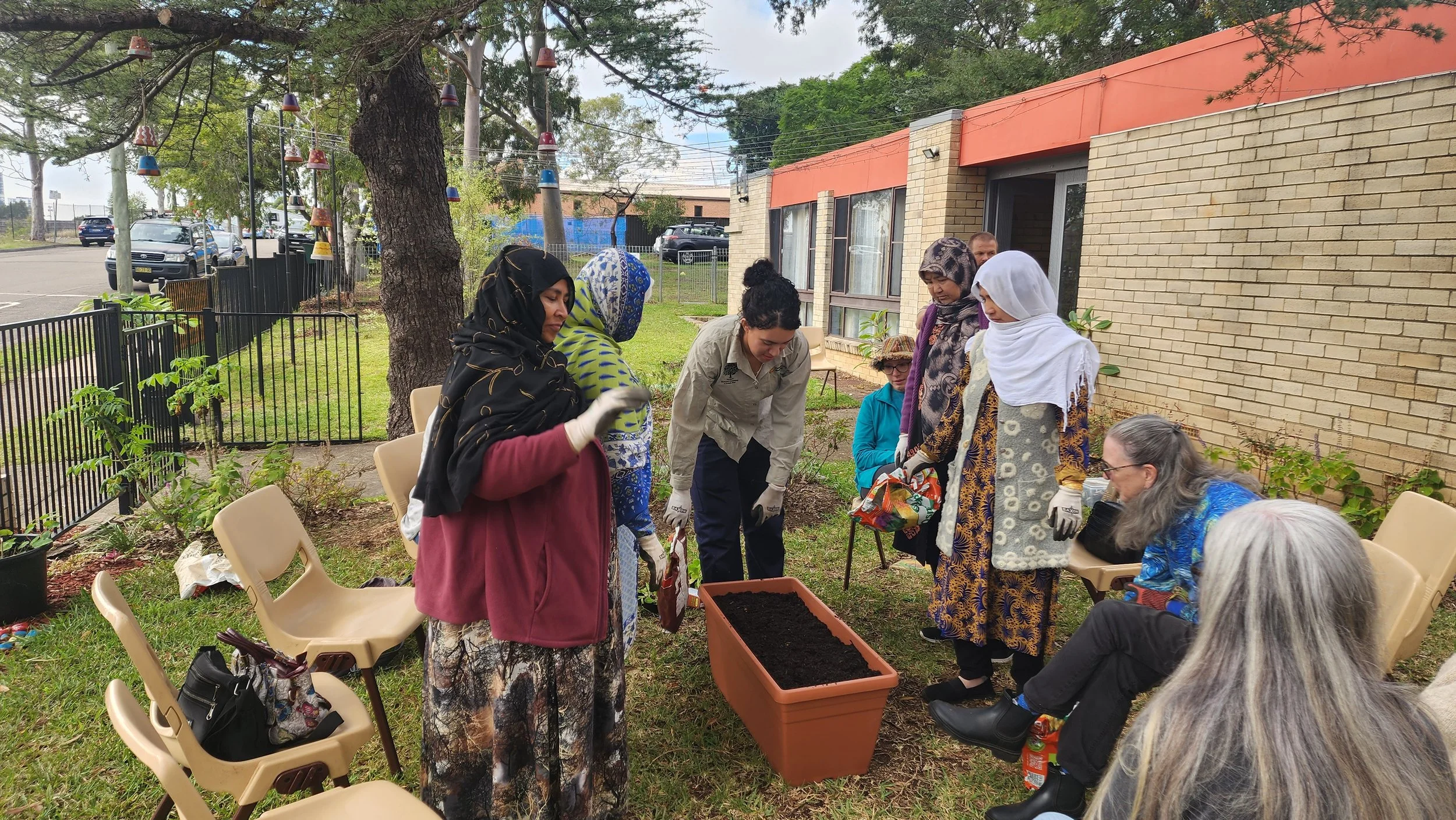 A group of diverse women gathered outdoors around a planter filled with dark soil. Some are standing, some sitting, engaged in a gardening activity, with a large tree and a brick building in the background.