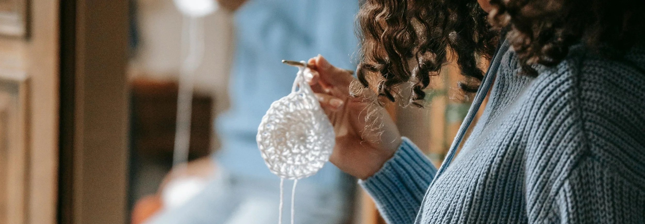 Person knitting with white yarn, wearing a blue sweater, curly hair, in a cozy indoor setting.