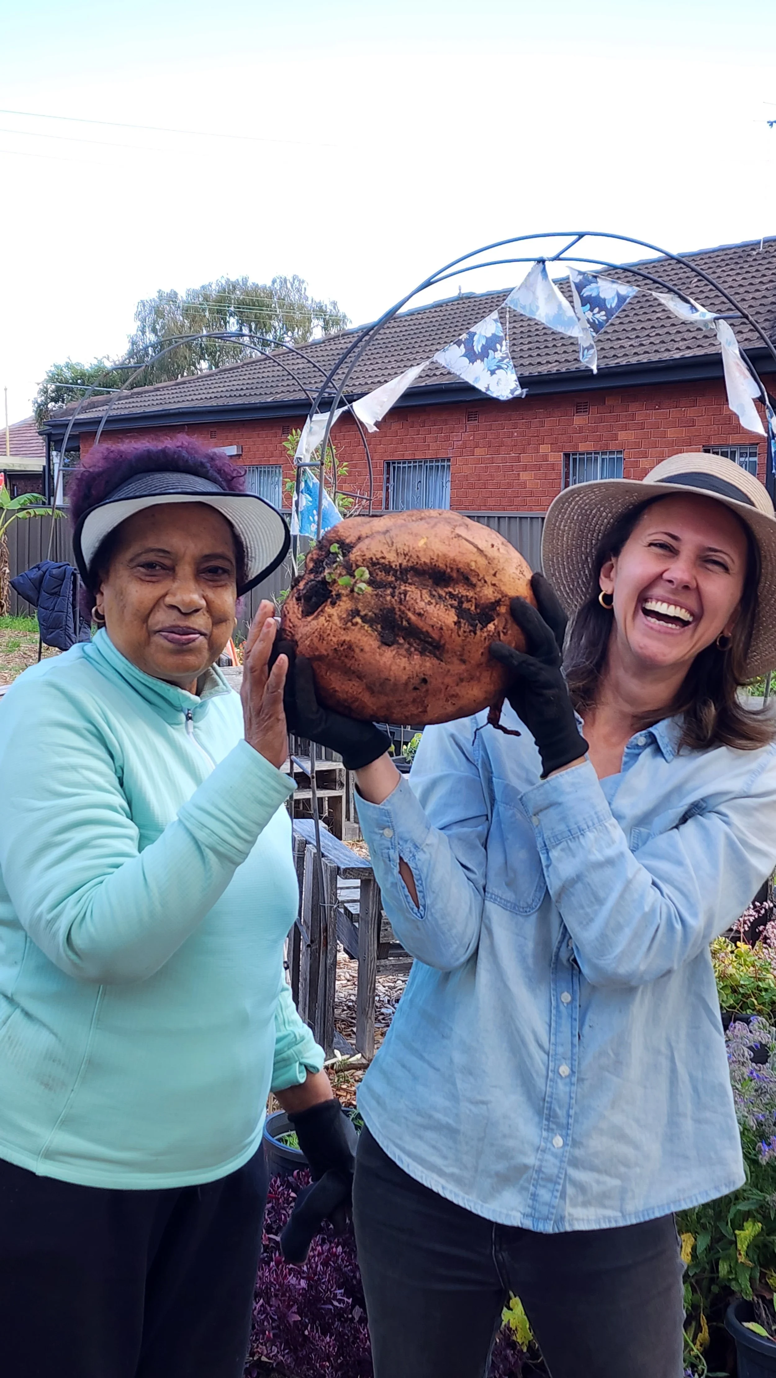 Two women smiling and holding a large, freshly harvested sweet potato in a garden with a red brick house and decorative bunting in the background.