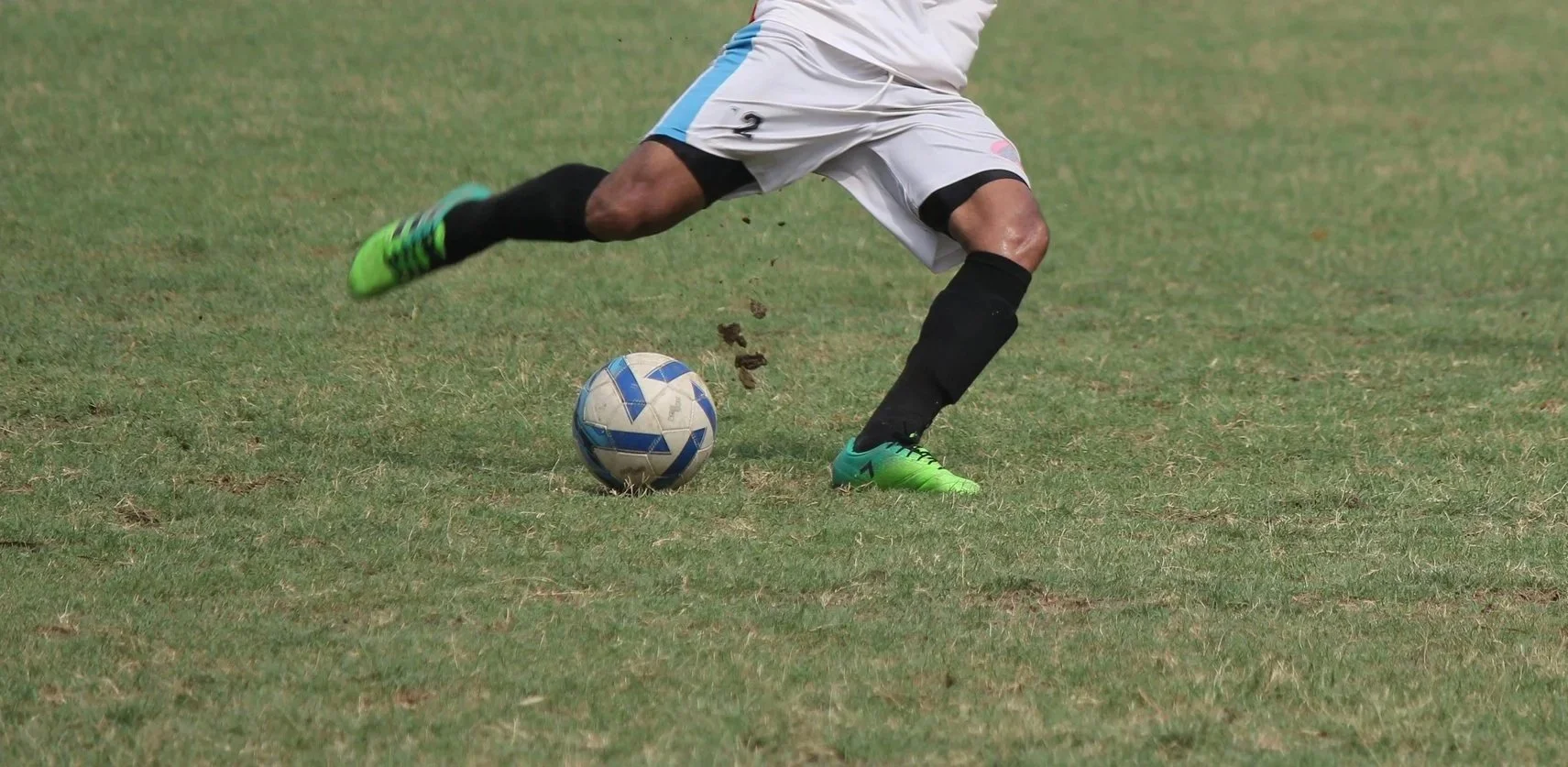 A soccer player in white and black uniform controls the ball on a grassy field, wearing green cleats.