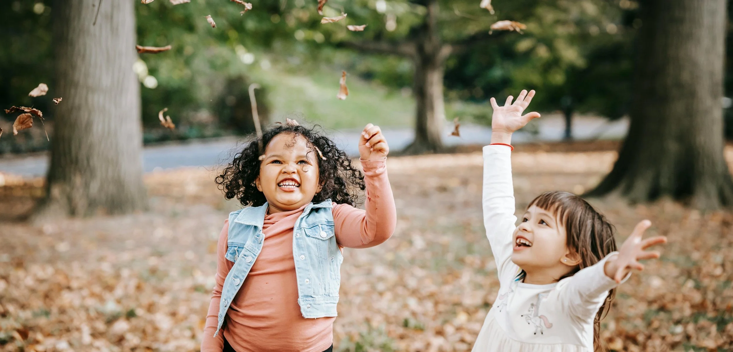 Two young girls playing and throwing fallen leaves in a forested park.