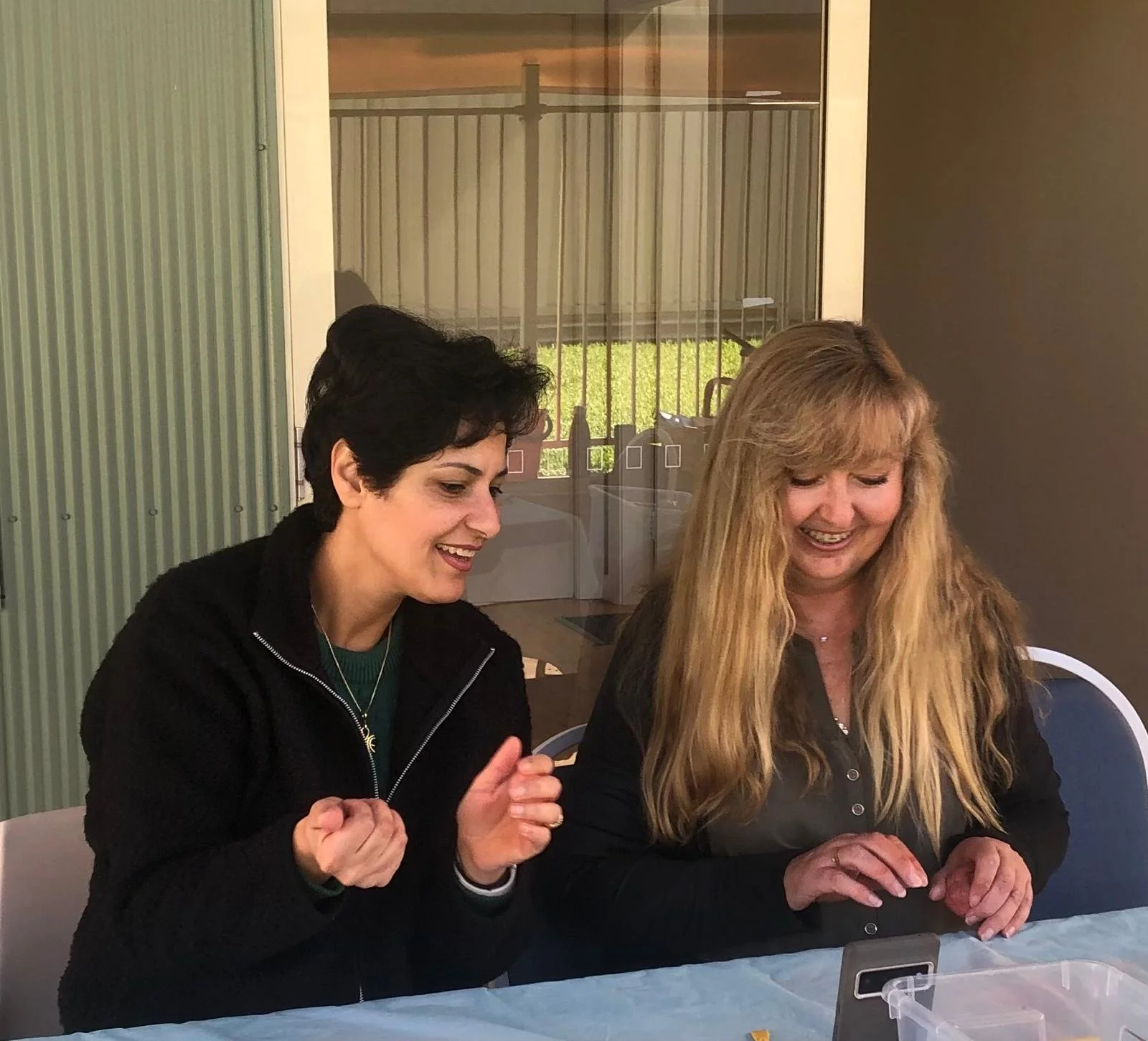 Two women sitting at a table, smiling and looking at a smartphone. One woman has short dark hair and is wearing a black jacket, the other has long blond hair and is wearing a dark blouse. There is a glass window with a sliding door behind them.