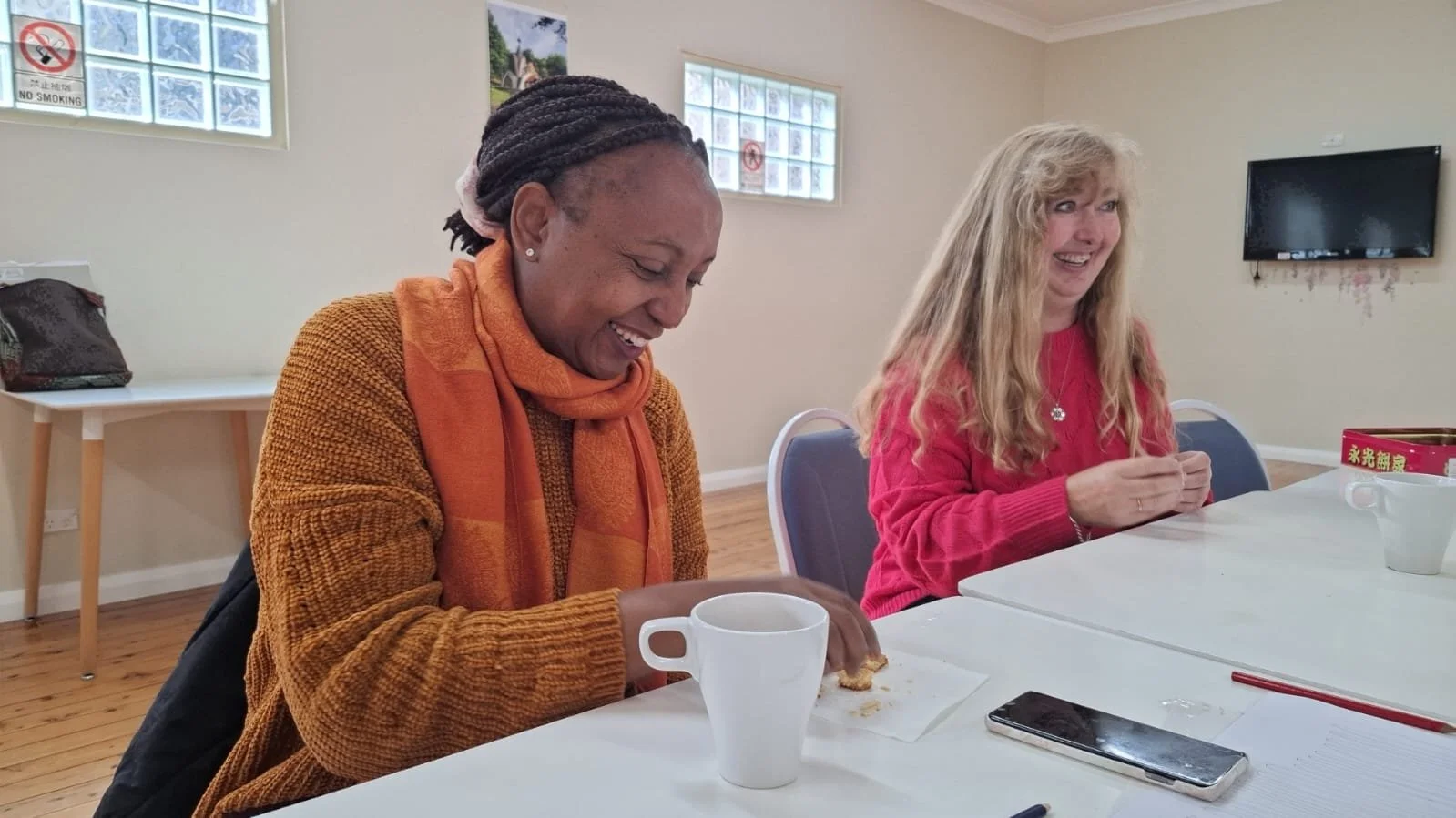 Two women sitting at a table, smiling and laughing. One woman with dark braided hair wearing an orange sweater and scarf, and the other with long blonde hair in a red sweater. There are cups, a smartphone, and papers on the table.