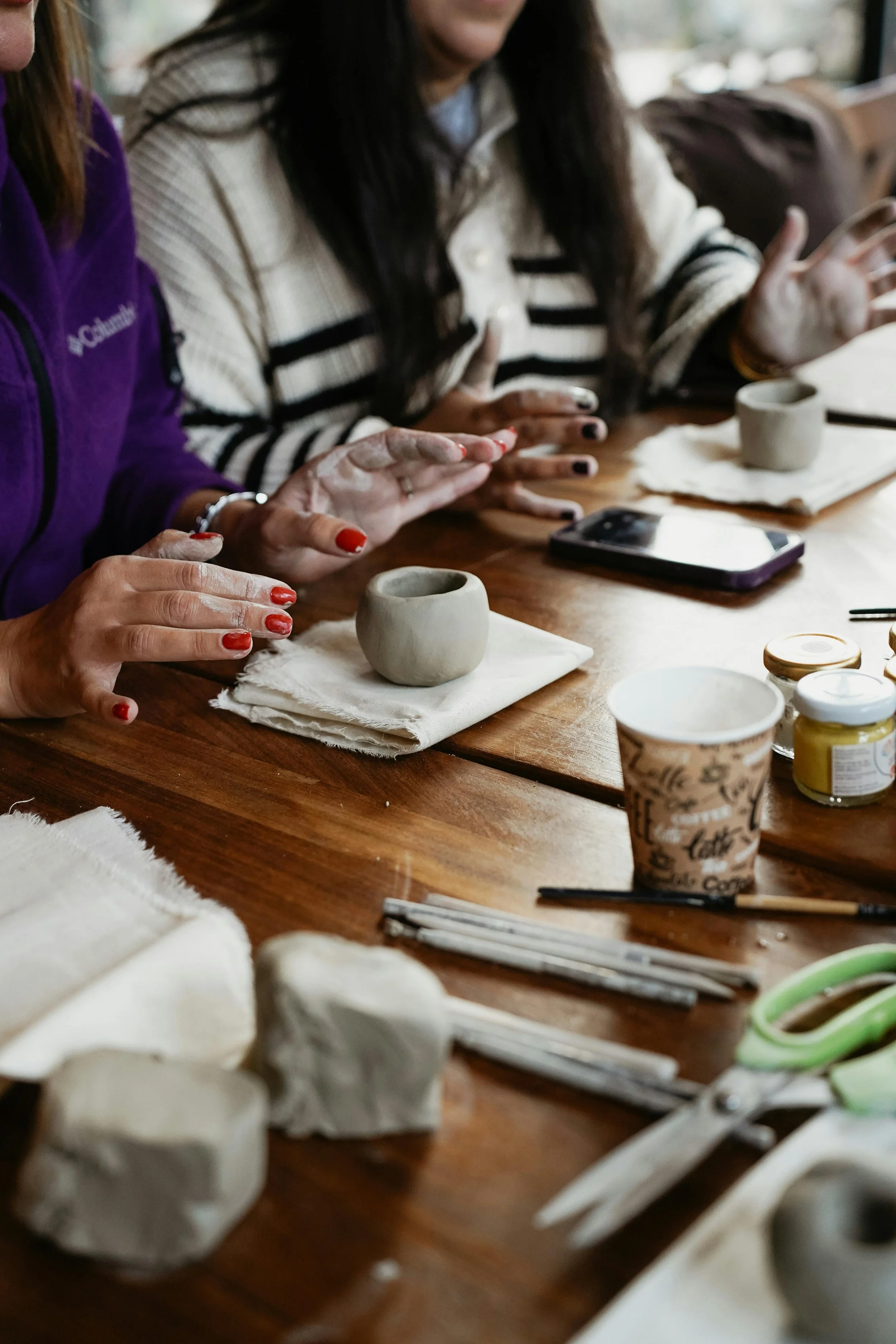 People working on pottery projects at a table with clay, tools, cups, and paint.