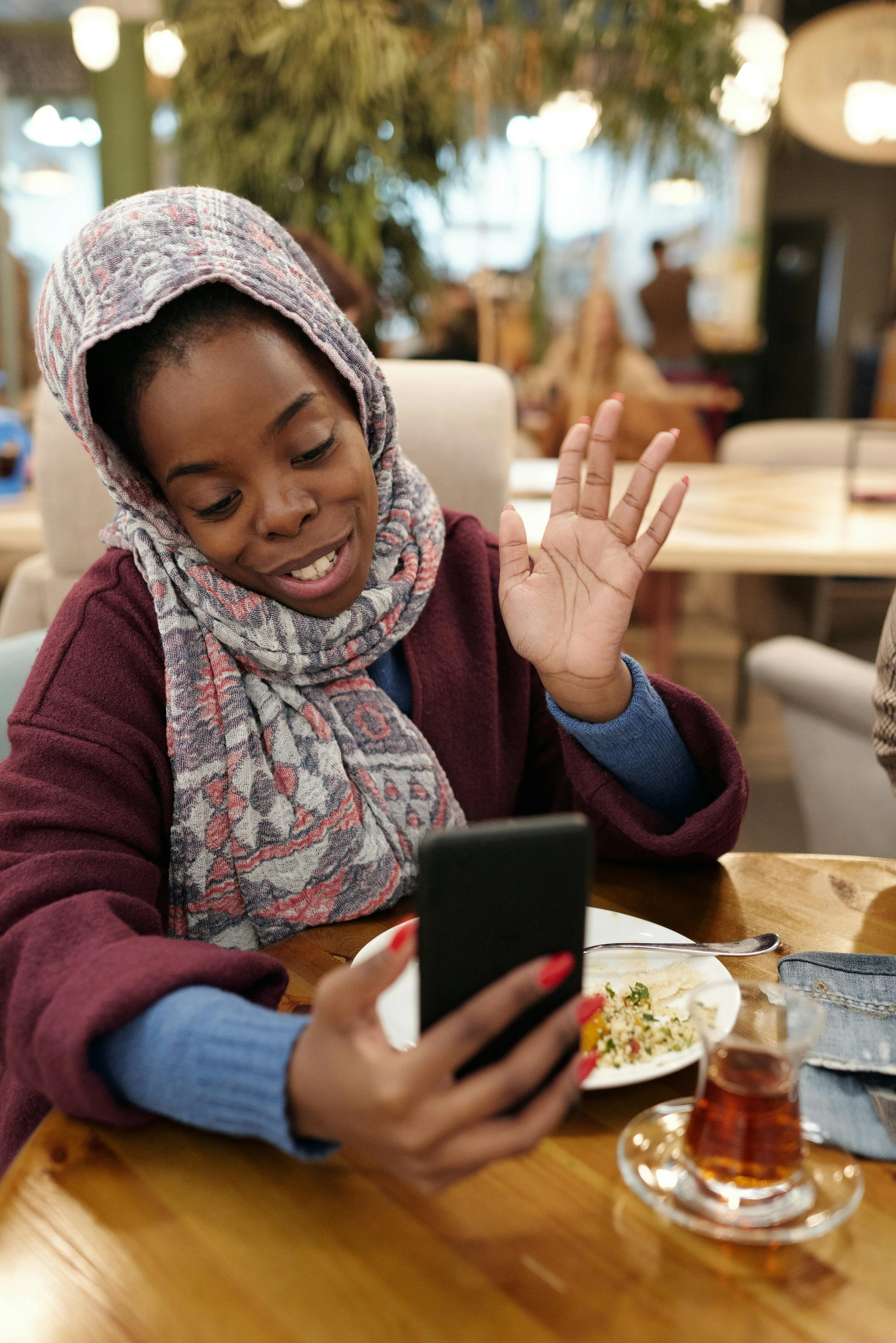 A woman wearing a colorful scarf and burgundy coat is sitting at a wooden table in a cozy restaurant, smiling and waving at her smartphone, with a plate of food, a glass of tea, and a denim napkin on the table.