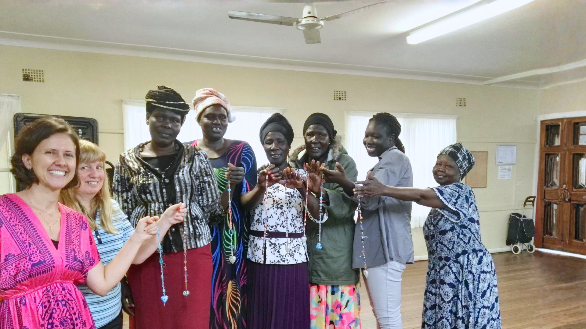 A group of women, including some of African descent and two of Caucasian descent, standing in a room with wooden flooring and light-colored walls. They are smiling and holding up beaded necklaces, with some women presenting or showing the jewelry. The women are dressed in a mix of casual and traditional clothing, and there is a ceiling fan above them and a wooden cabinet on the right side of the room.
